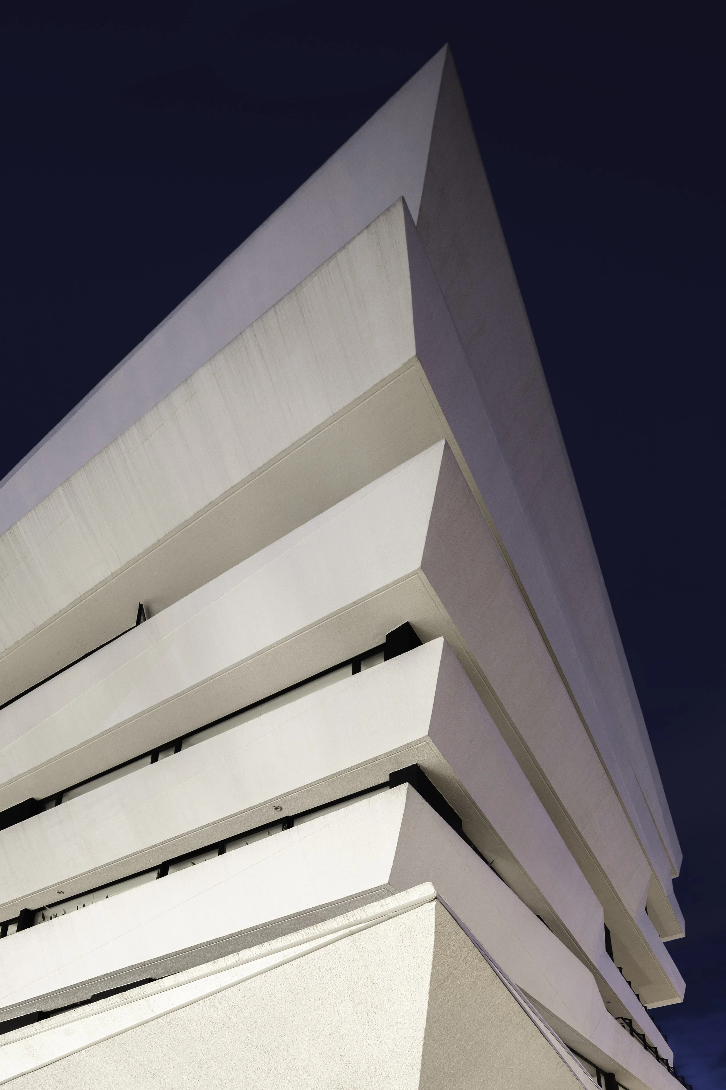 Vista desde abajo de un edificio moderno de color blanco con varias terrazas y barandales, con cielo azul oscuro en el fondo.