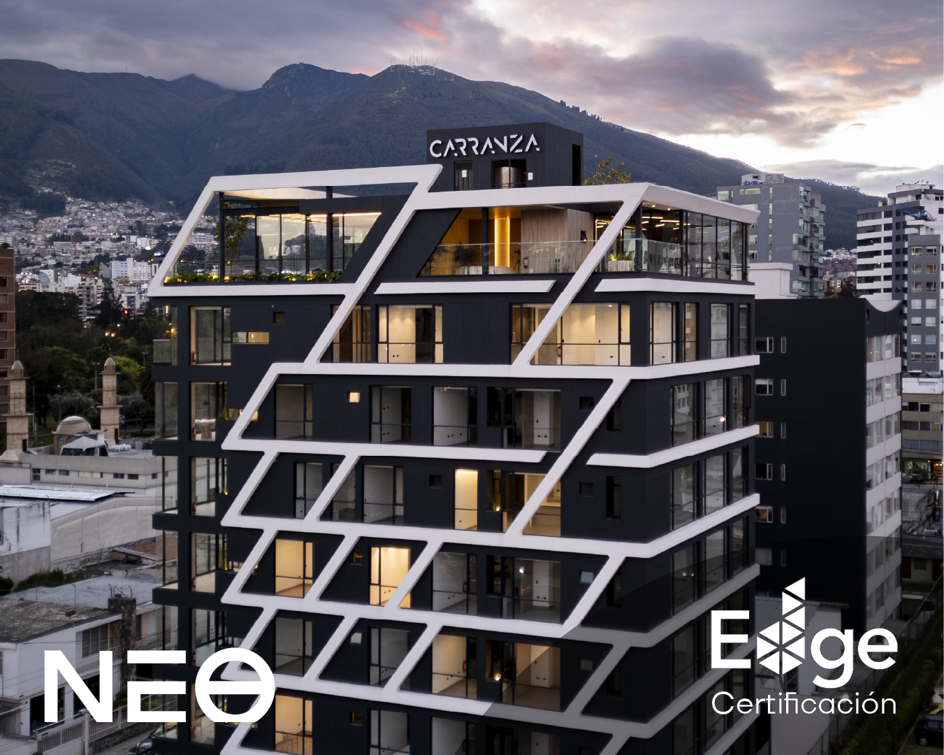 Edificio moderno de varios pisos con estructura blanca en forma de línea irregular, con ventanas grandes y balcones, en una ciudad con montañas de fondo y un cielo al atardecer.