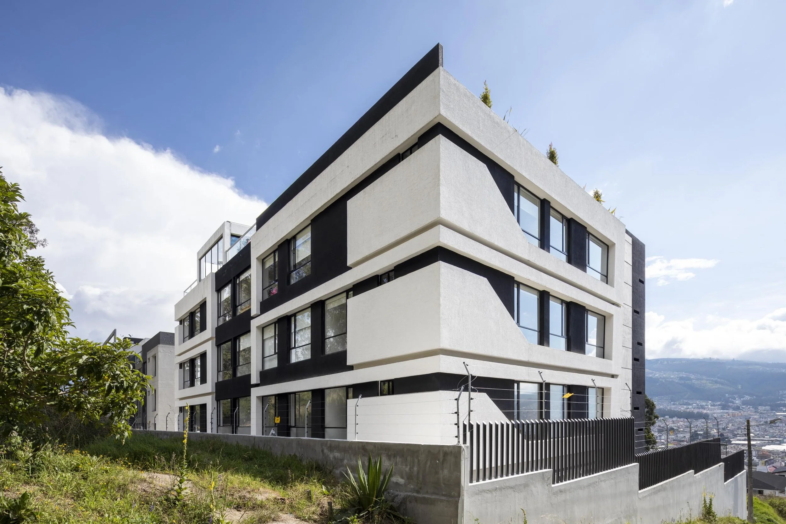 Edificio moderno de varios pisos con ventanas grandes y balcones, rodeado de naturaleza y con vista a una ciudad y montañas al fondo.