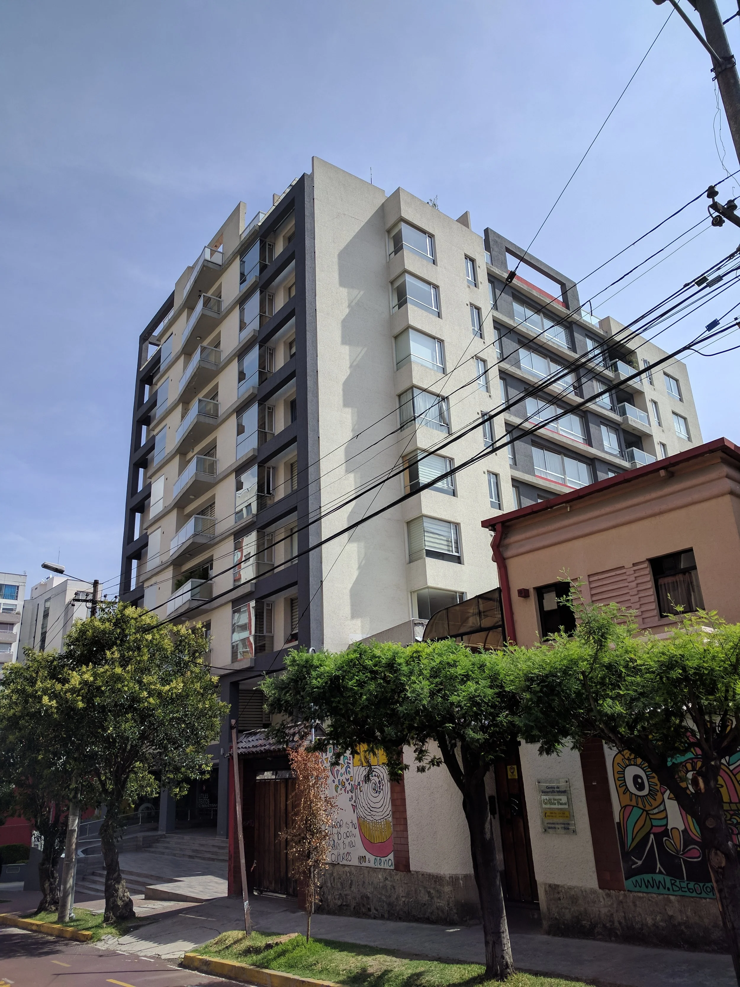 Edificio de varios pisos con balcones y ventanas, árboles en la acera y murales en la pared de la entrada, en un día soleado.