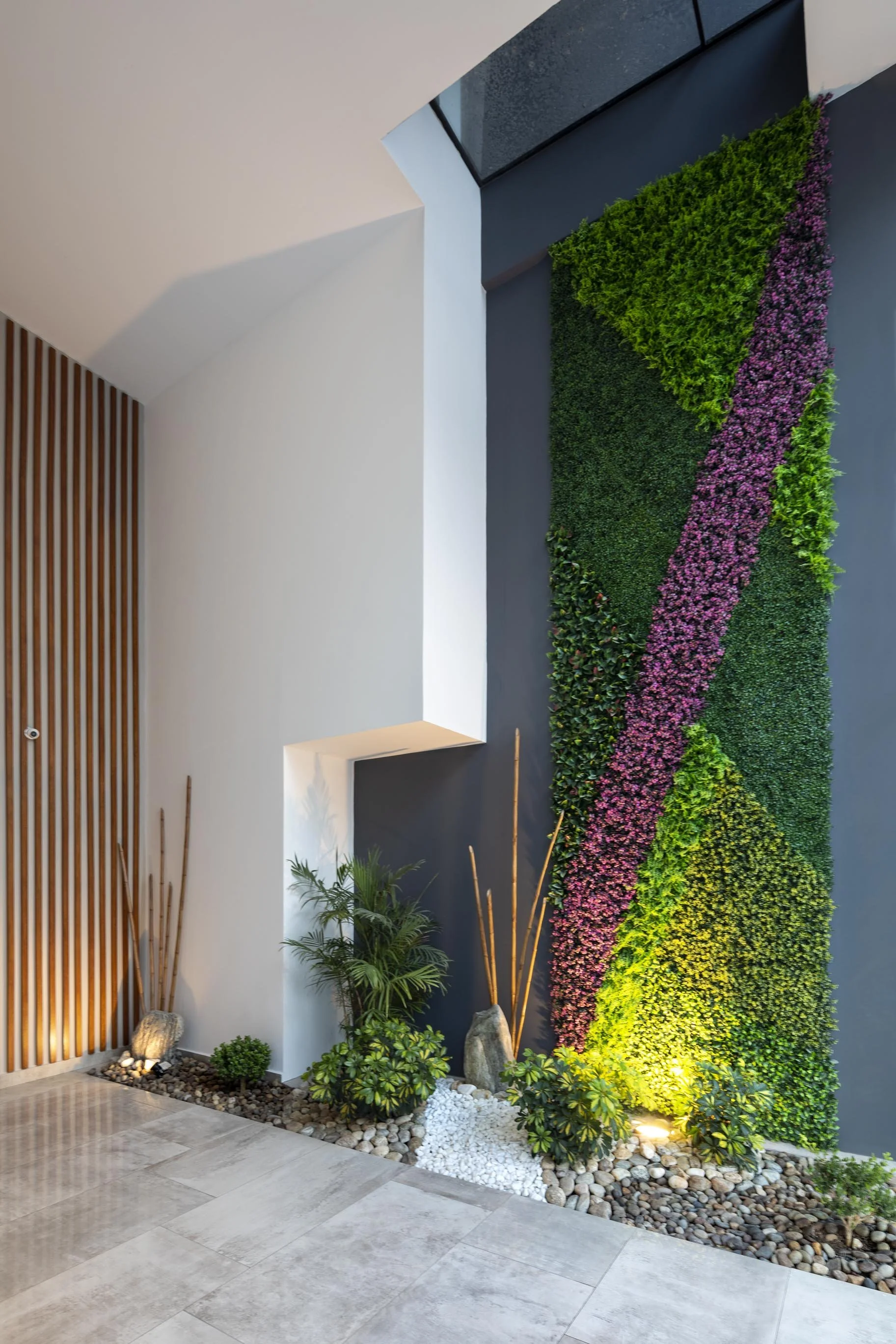 Pared con muro vegetal decorativo con plantas verdes y flores rosas, iluminación artificial que resalta la vegetación, piso de baldosas claras y pared blanca de fondo.