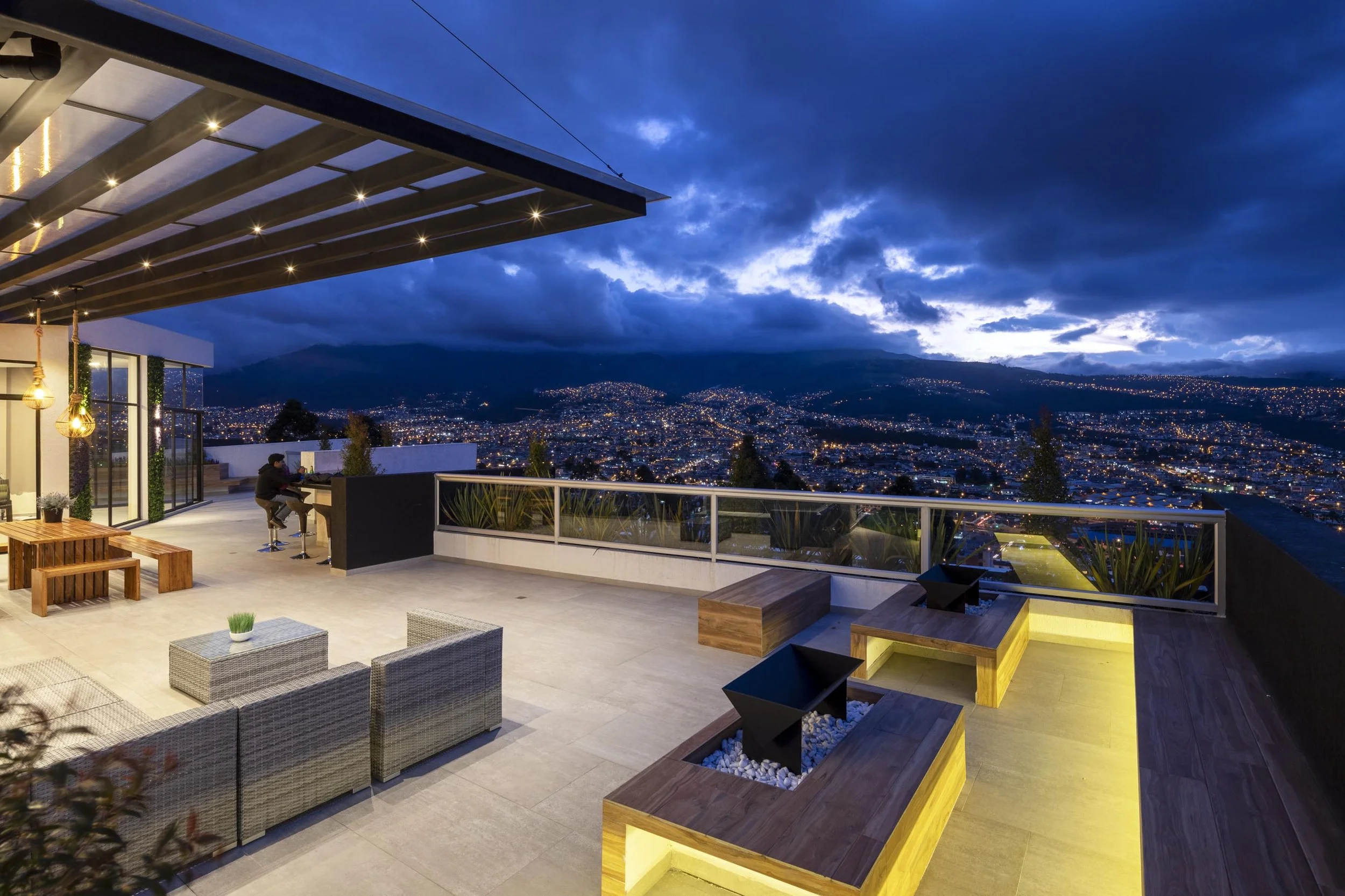 Terraza moderna con vista nocturna de la ciudad, con sillones, mesas de madera, plantas y un piano, iluminada suavemente, en una noche lluviosa.