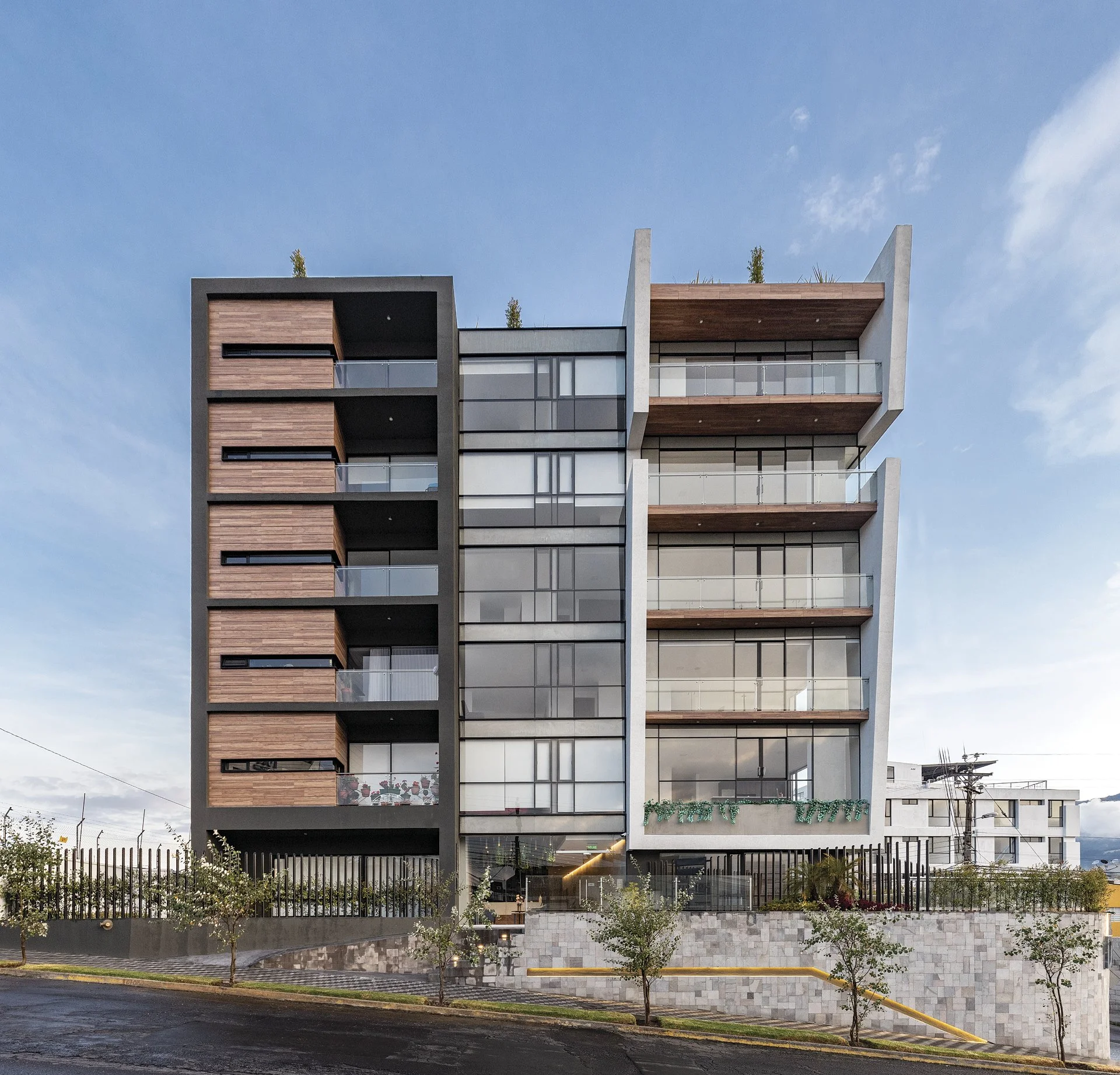 Edificio moderno de varias plantas con fachada de vidrio y madera, en una calle con árboles y cielo despejado.