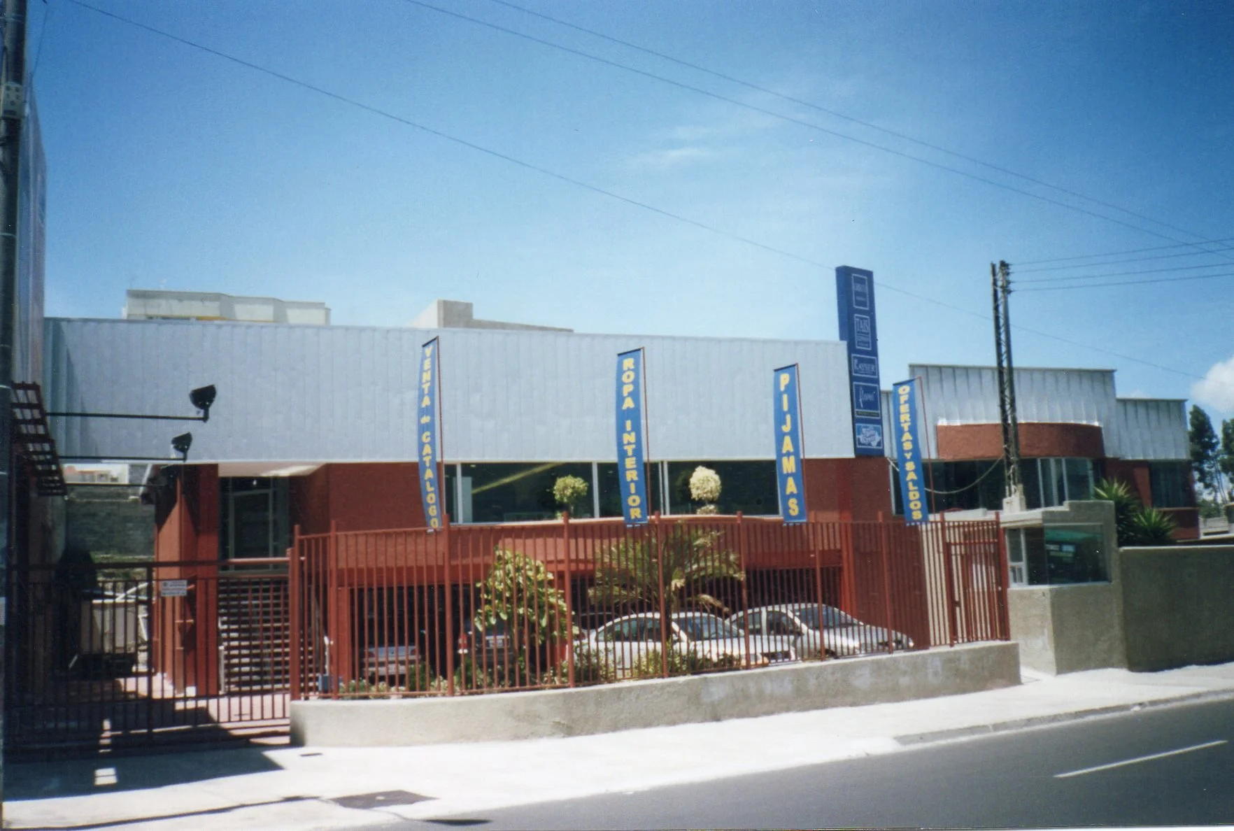 Edificio de color rojo con señalización azul y blanca que anuncia libros y otros servicios en letras grandes, con un estacionamiento frente y una reja roja.