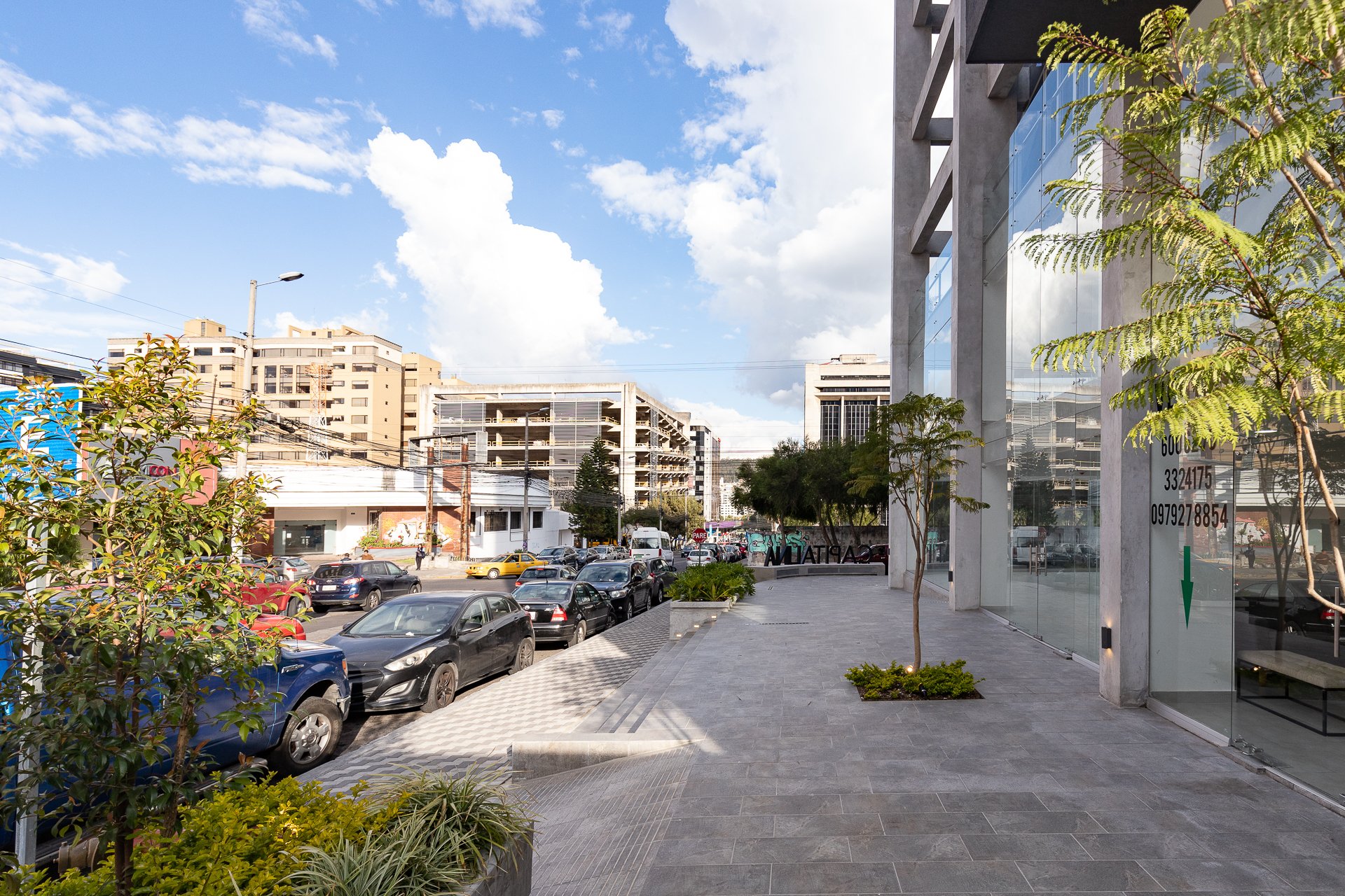 Una vista de la ciudad con edificios modernos, árboles, autos estacionados y una acera amplia bajo un cielo despejado con algunas nubes.