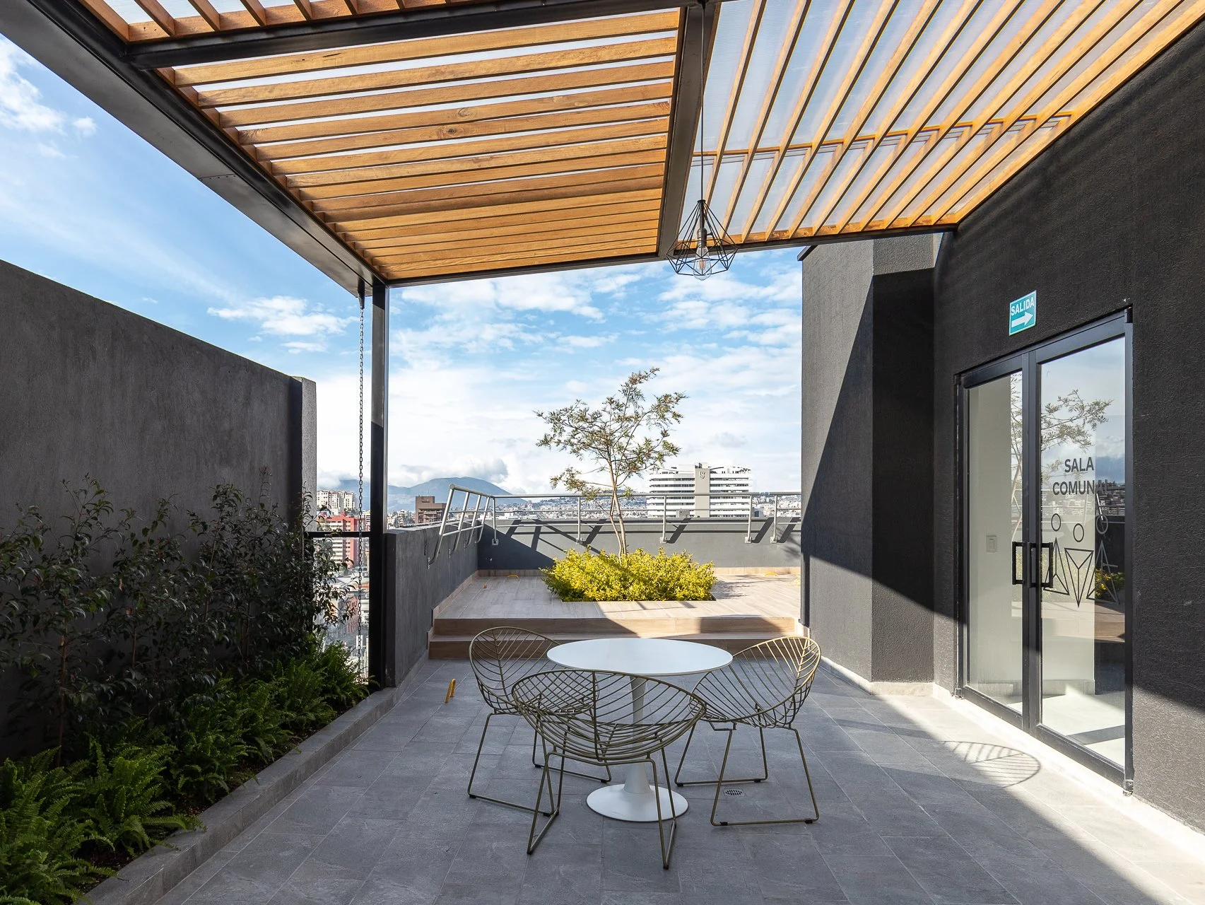 Terraza moderna con sillas de alambre y una mesa redonda, zona de plantas verdes y un árbol pequeño, con vista a la ciudad y un cielo despejado.