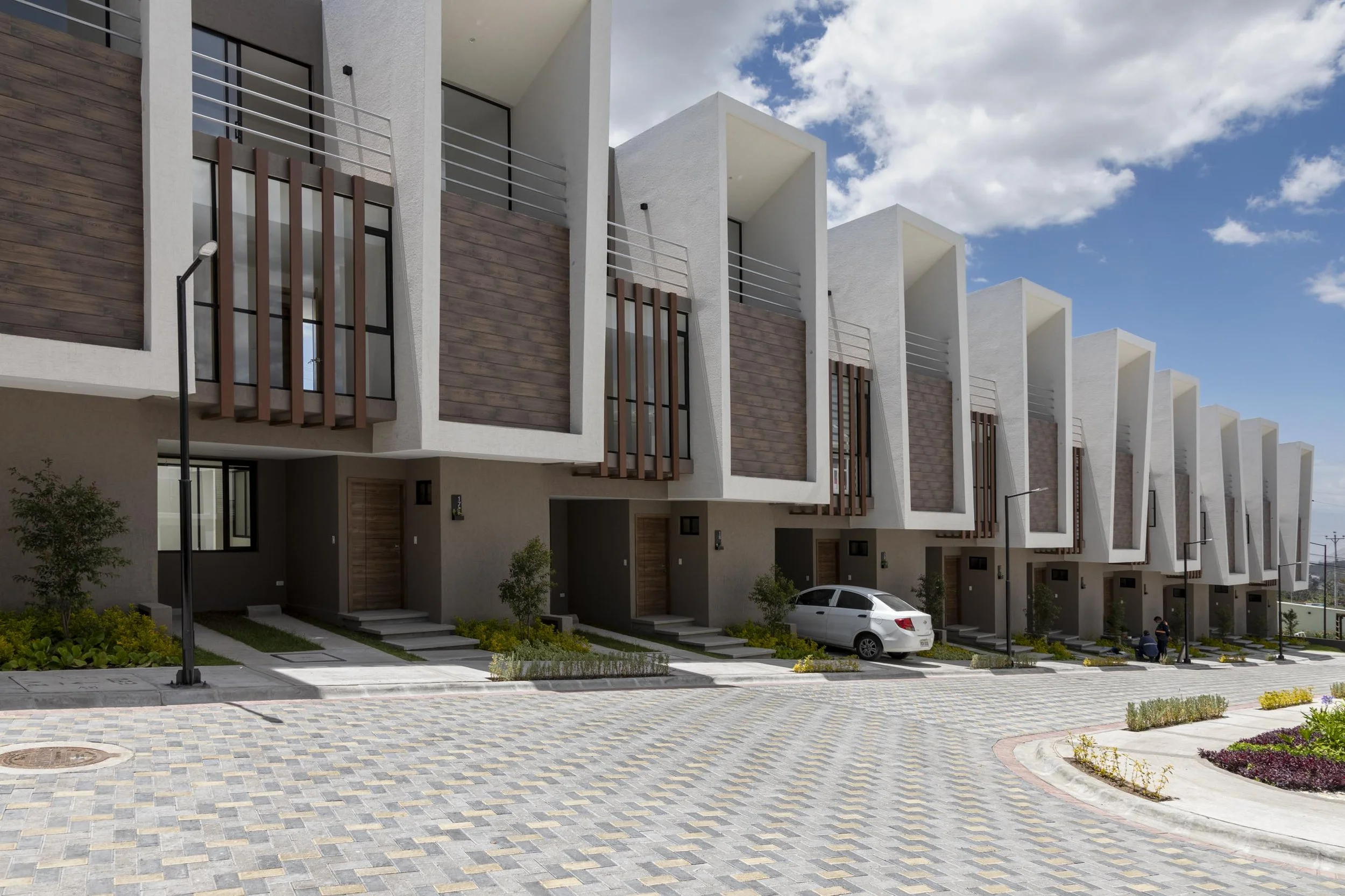 Conjunto de casas modernas de varias plantas, con fachadas de colores claros, barandillas de metal, y estacionamiento en la calle, bajo un cielo parcialmente nublado.