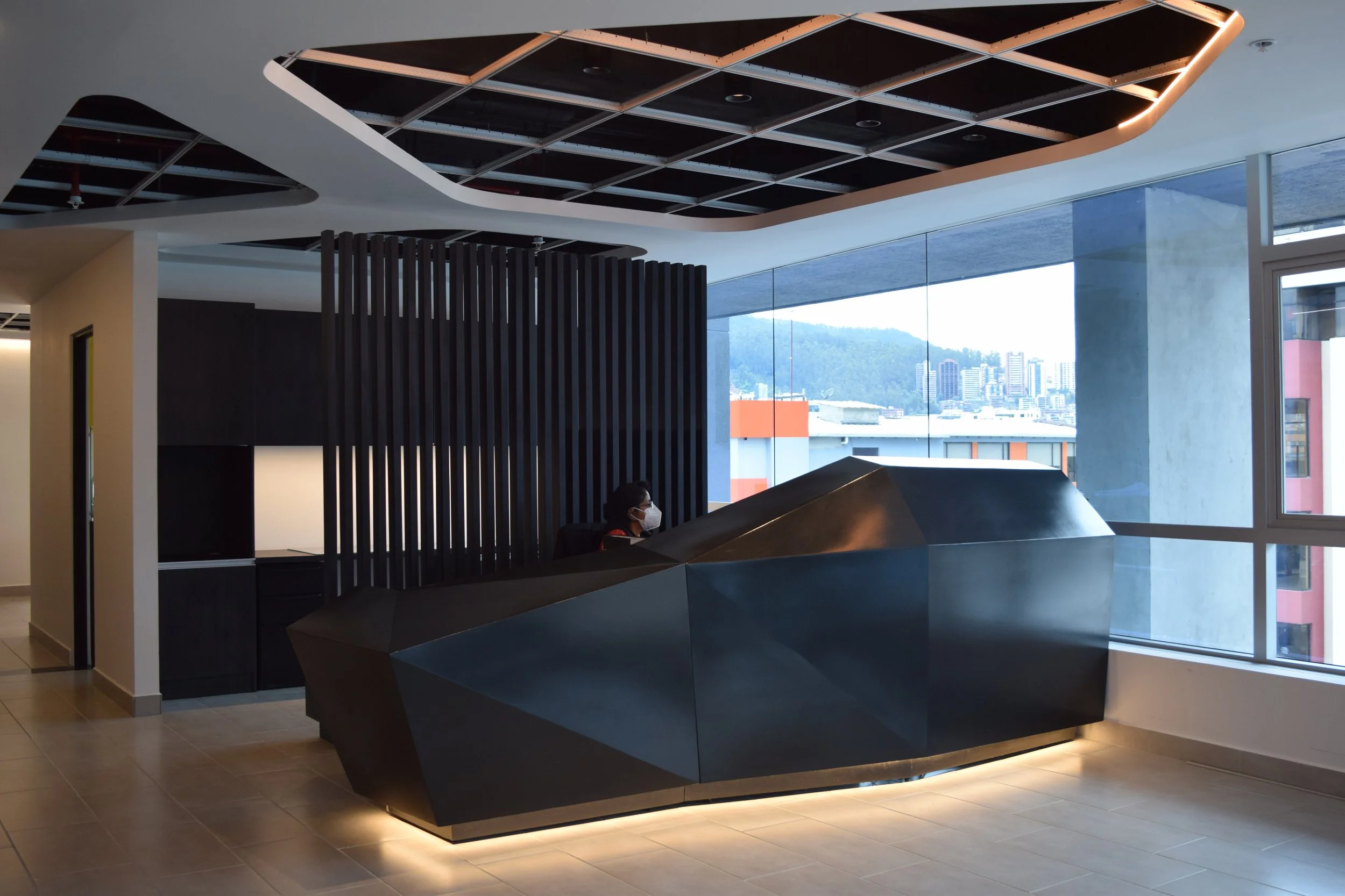 Recepcionista con mascarilla en una recepción moderna de edificio, con vista a la ciudad y montañas a través de ventanales.