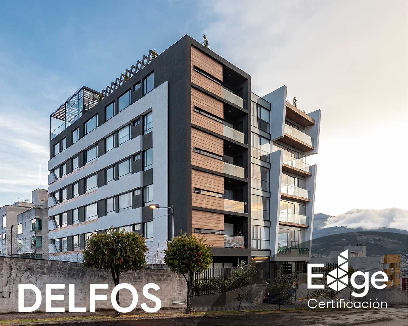 Edificio moderno de varios pisos con balcones de vidrio y fachada de madera y gris, con montañas al fondo y árbol en primer plano.