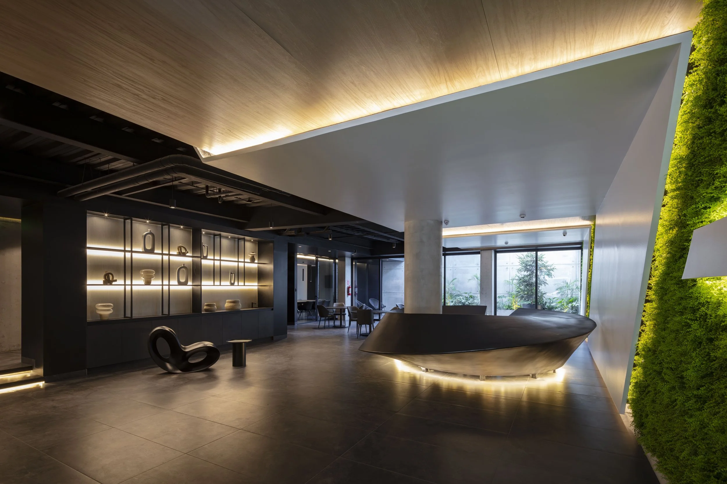 Interior de edificio moderno en quito ecuador con paredes blancas, piso de concreto, luz ambiental cálida, muebles de madera y acrílicos, y vegetación de paredes verdes a la derecha.