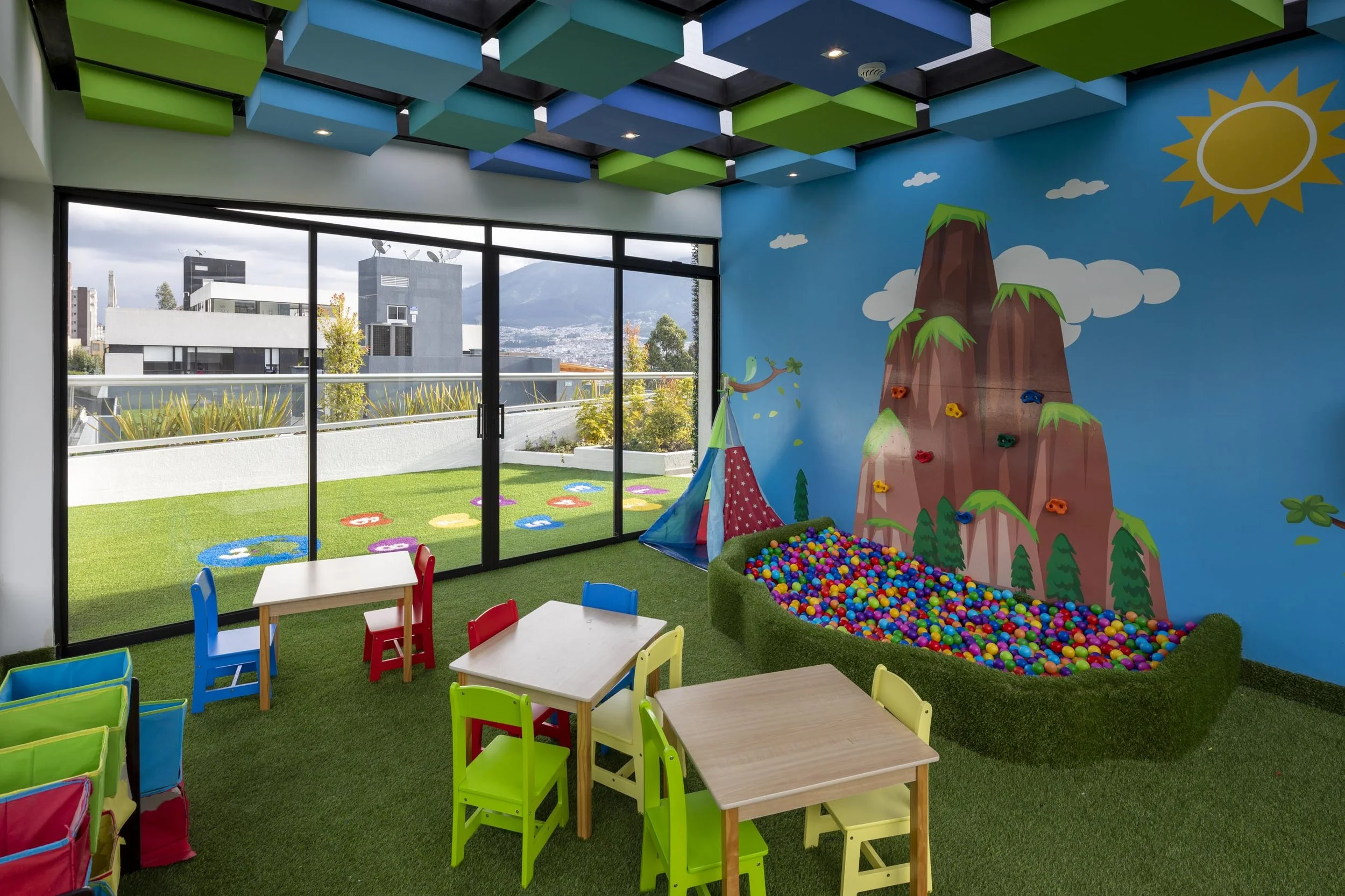 Sala de juegos infantiles con paredes decoradas, torre de rocas, piscina de bolas y mesas con sillas coloridas, vista a un balcón con césped artificial y paisaje urbano.