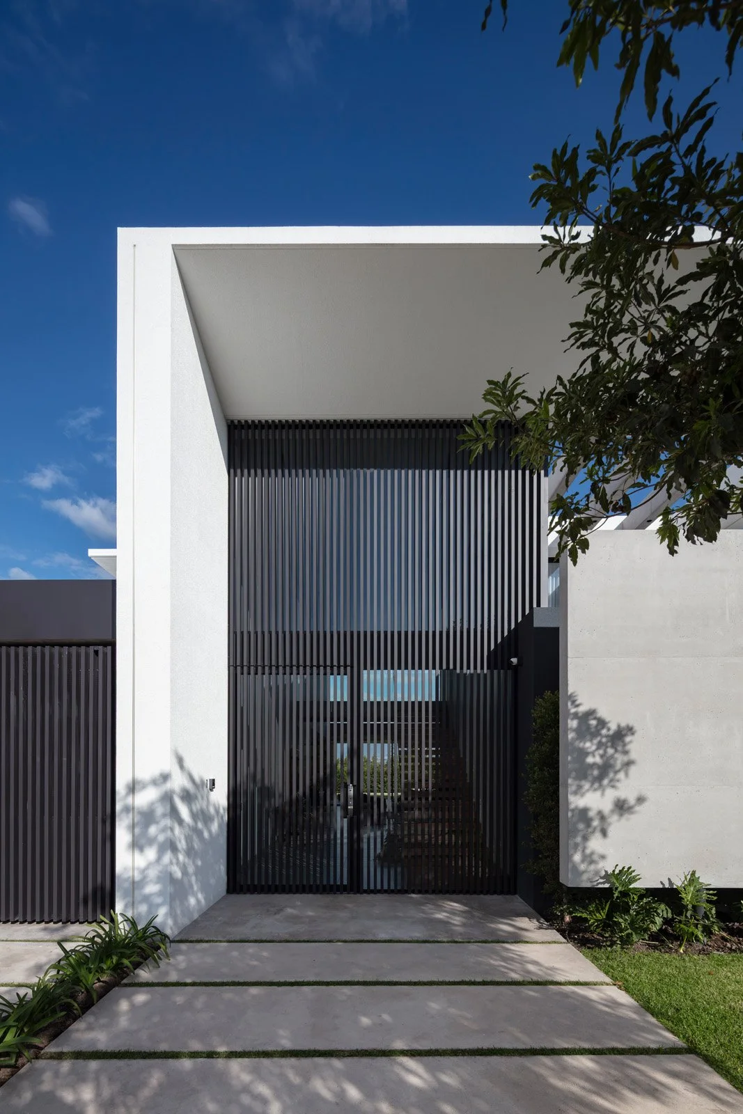 Entrada moderna de una casa con rejas negras verticales y muro blanco, rodeada de vegetación y camino de concreto.