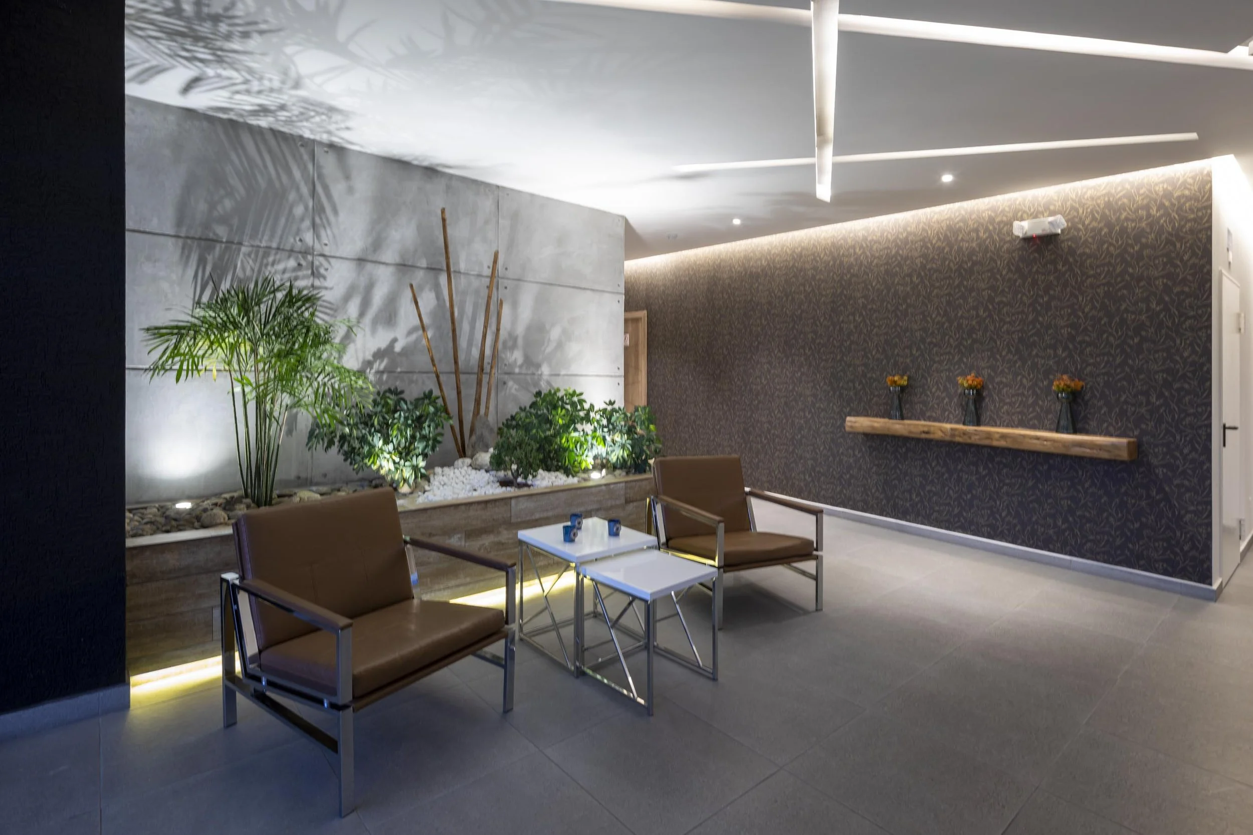 Sala de espera con dos sillas de cuero marrón, mesas blancas, plantas verdes y decoración moderna en paredes gris y con papel tapiz. 