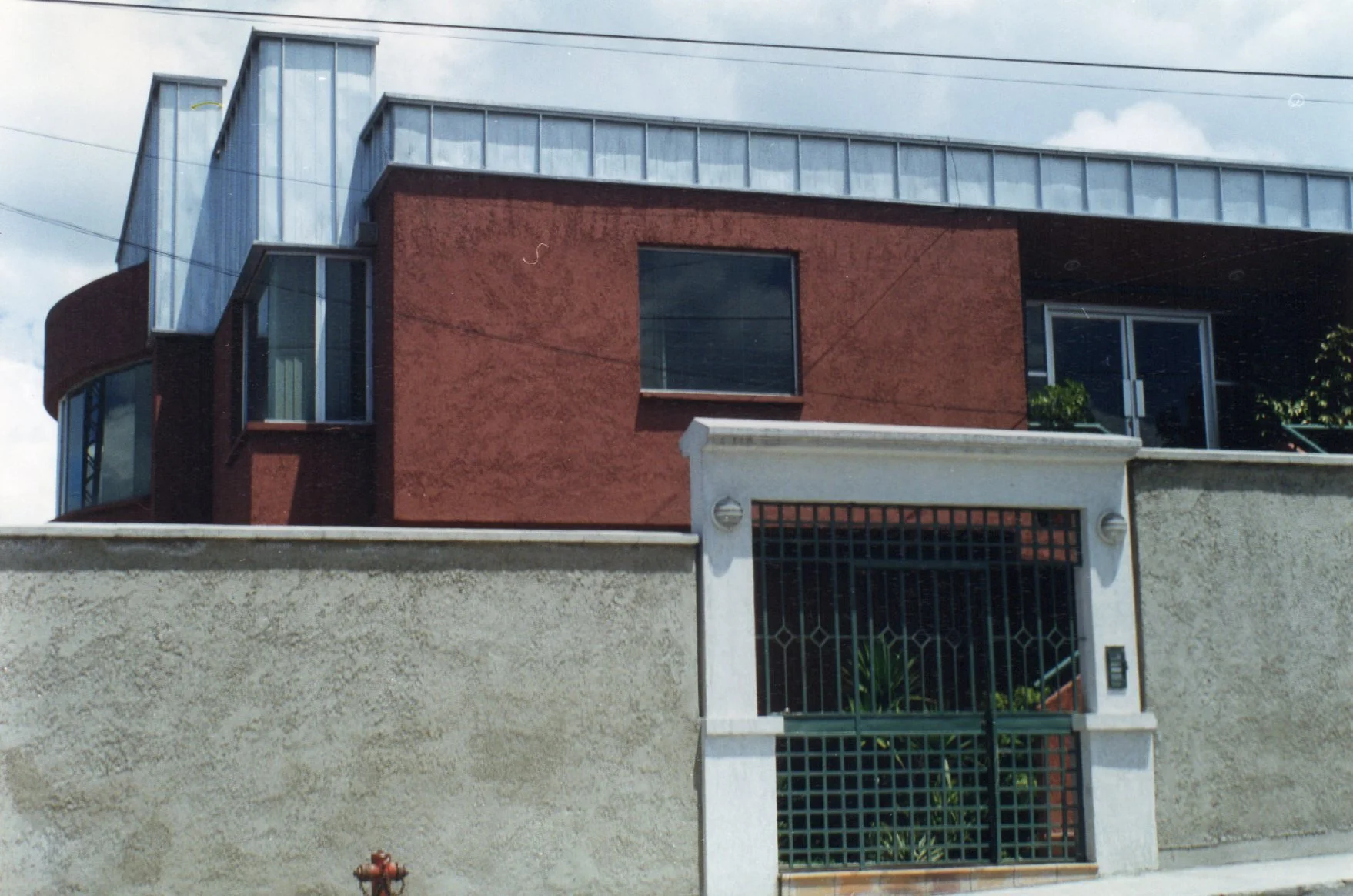 Casa moderna de varias plantas con muro exterior de color rojo, ventanas grandes de vidrio y estructura moderna en la parte superior, con un muro de seguridad en la entrada.