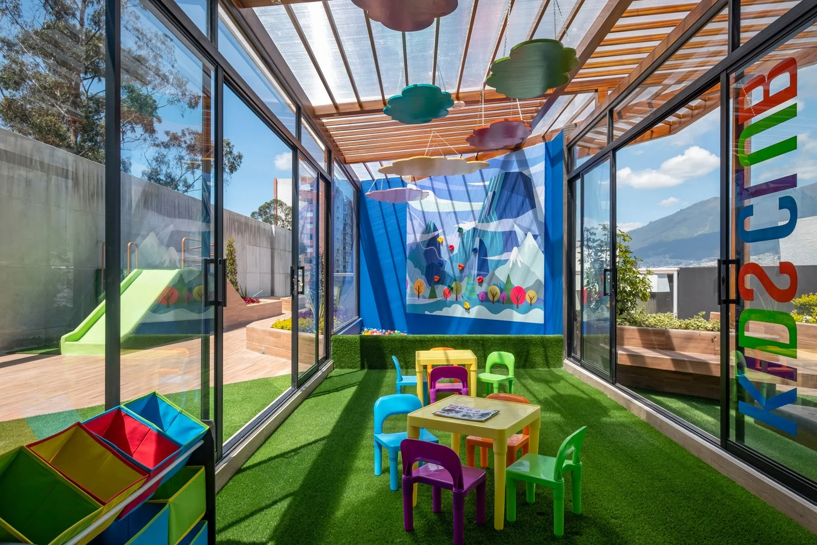 Un área de juegos para niños con paredes de vidrio y césped artificial. Incluye mesas y sillas de colores, un tobogán verde, y una pared decorativa con montañas y árboles. Hay nubes decorativas colgando del techo de madera.