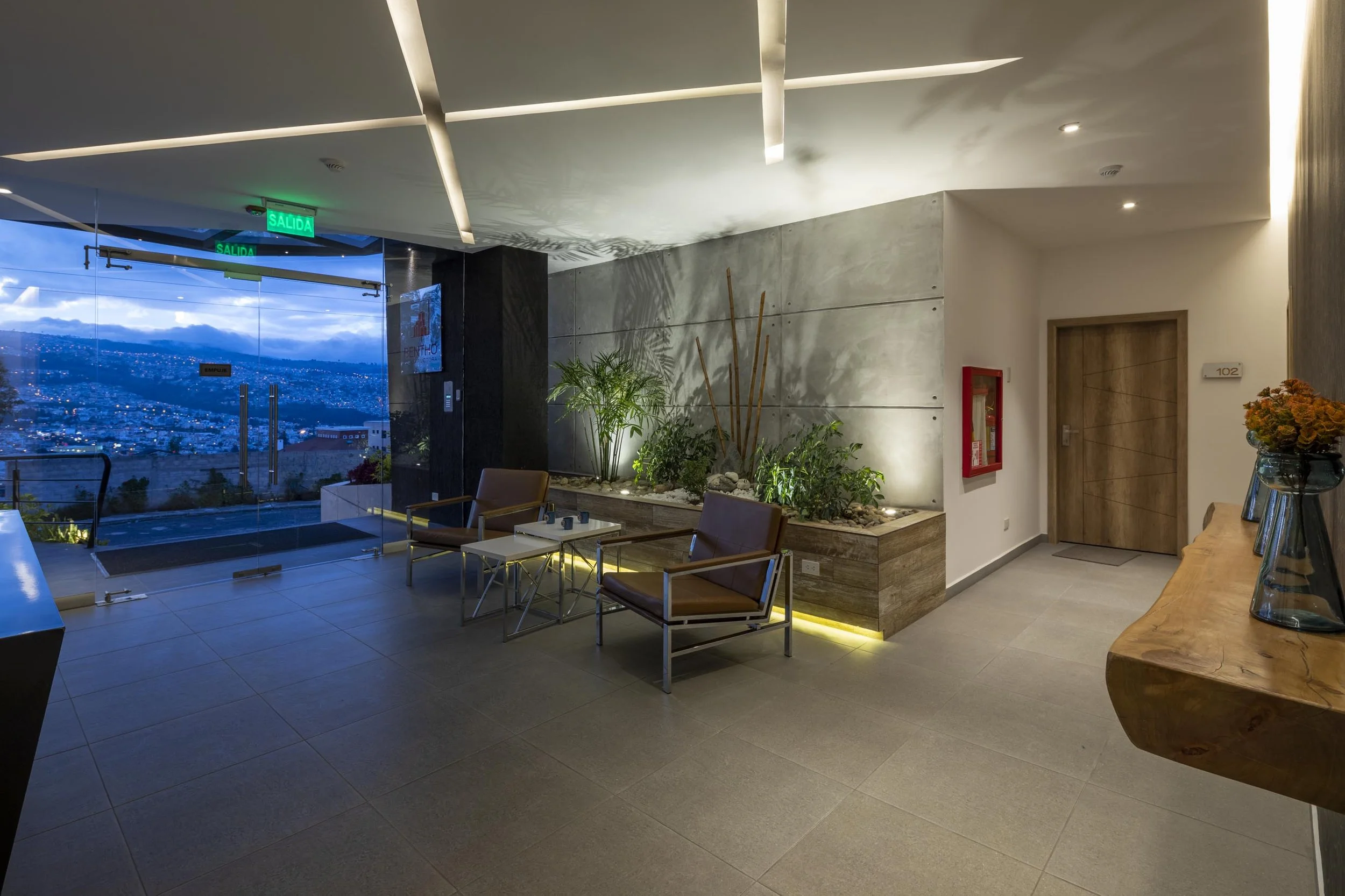 Vestíbulo de un edificio con puertas de vidrio, plantas decorativas y sillones de descanso, con vista a una ciudad y colinas al atardecer.