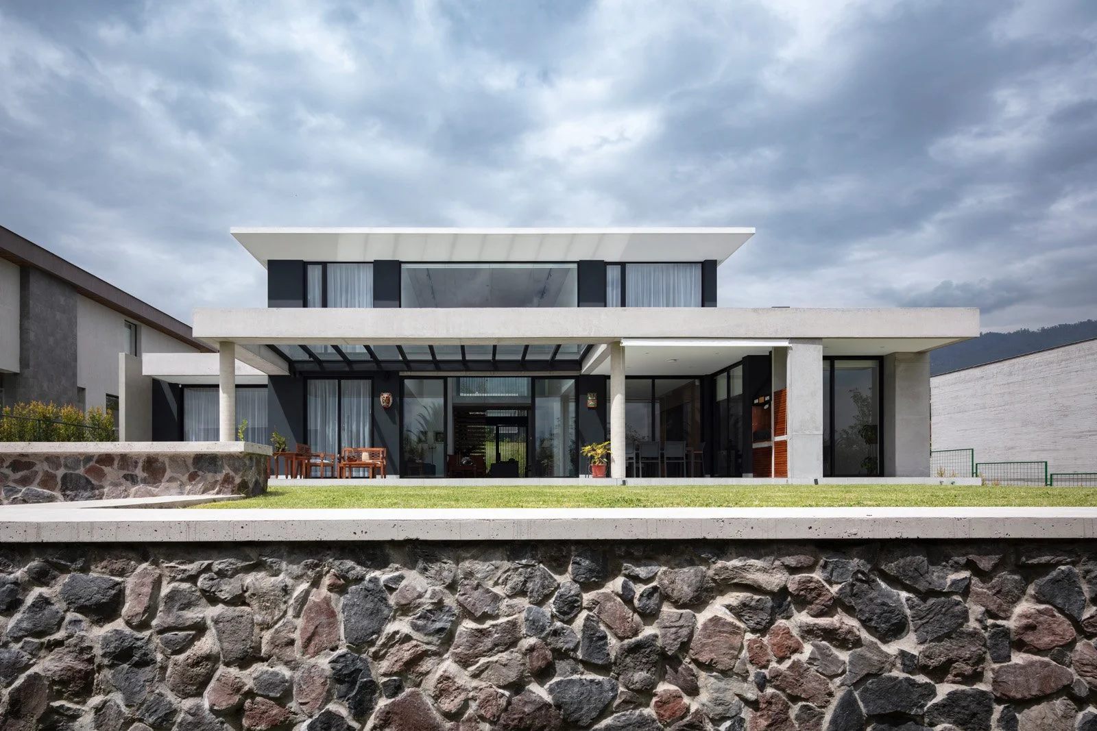 Casa moderna de varios niveles con grandes ventanas, terraza y jardines, bajo un cielo nublado.