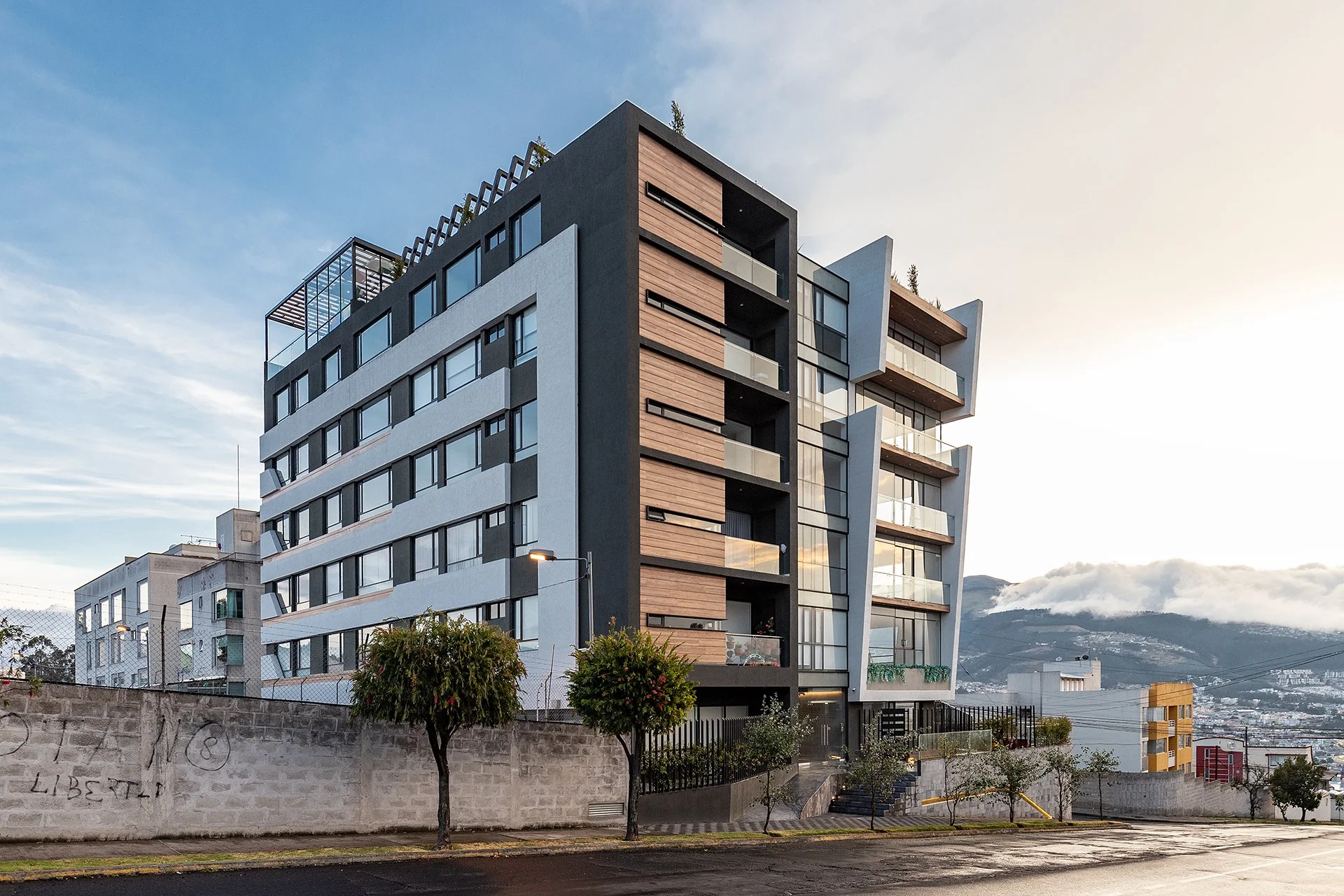 Edificio moderno de varios pisos con balcones de cristal y paneles de madera, en un fondo de montaña y cielo despejado, con algunas plantas y árboles en la calle frente al edificio.
