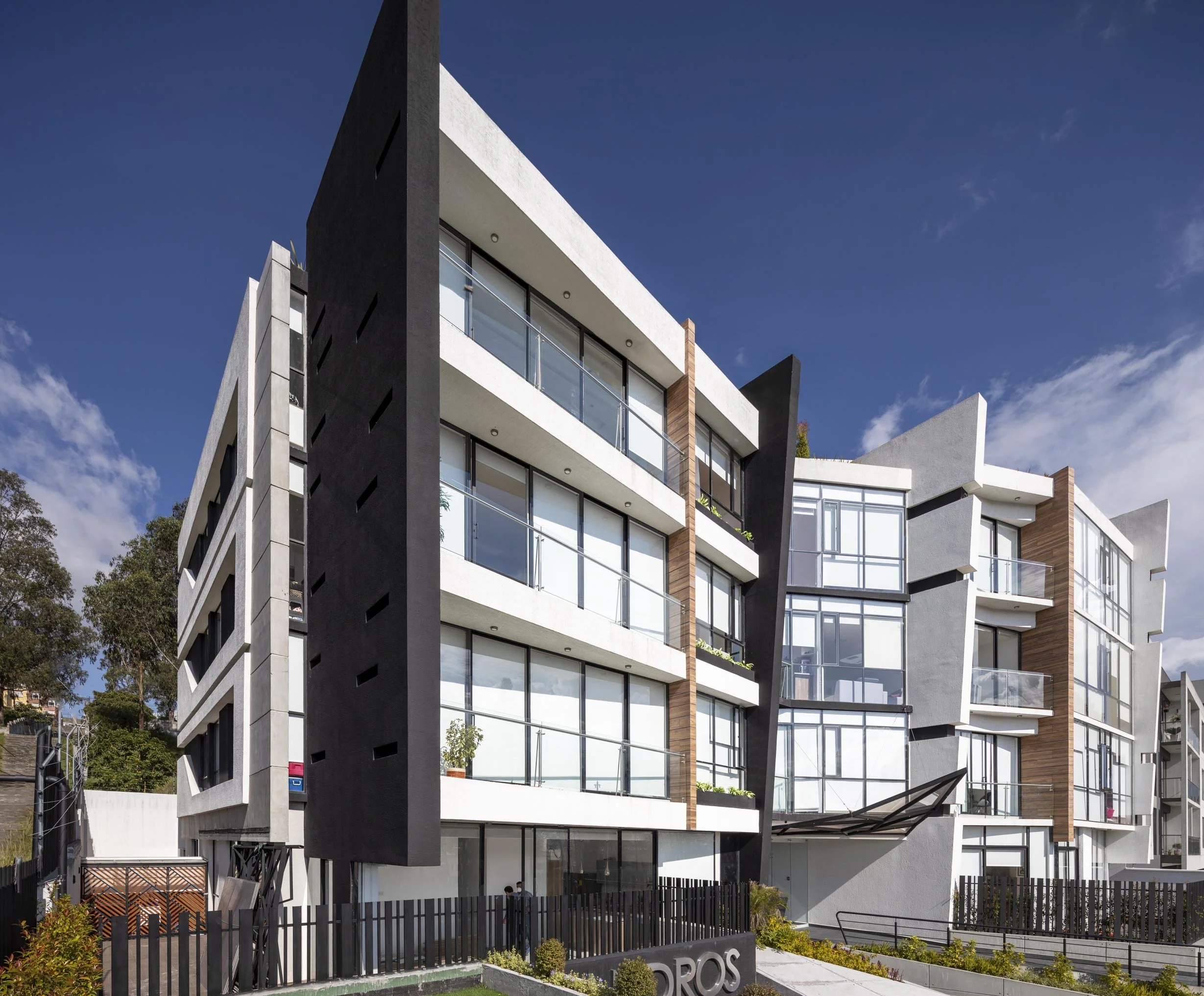 Edificio moderno de varios pisos con fachada de ventanas de vidrio y paredes blancas y negras, bajo un cielo azul con algunas nubes.