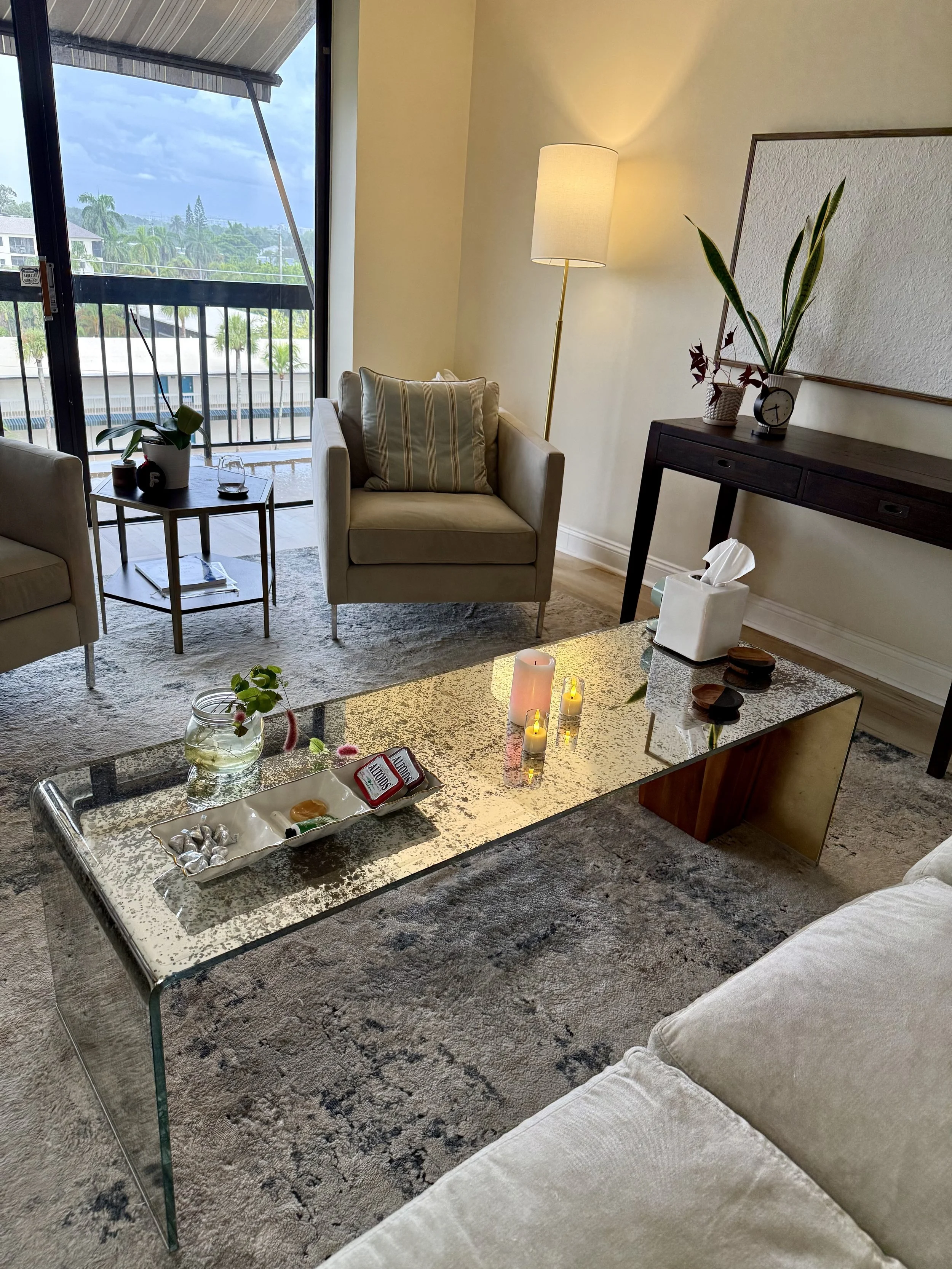 Living room with beige sofa, glass coffee table with decorative items and candles, side table with potted plants and glass of water, a tall floor lamp, and a wooden console table with a clock and plants. Large window with balcony and city view.