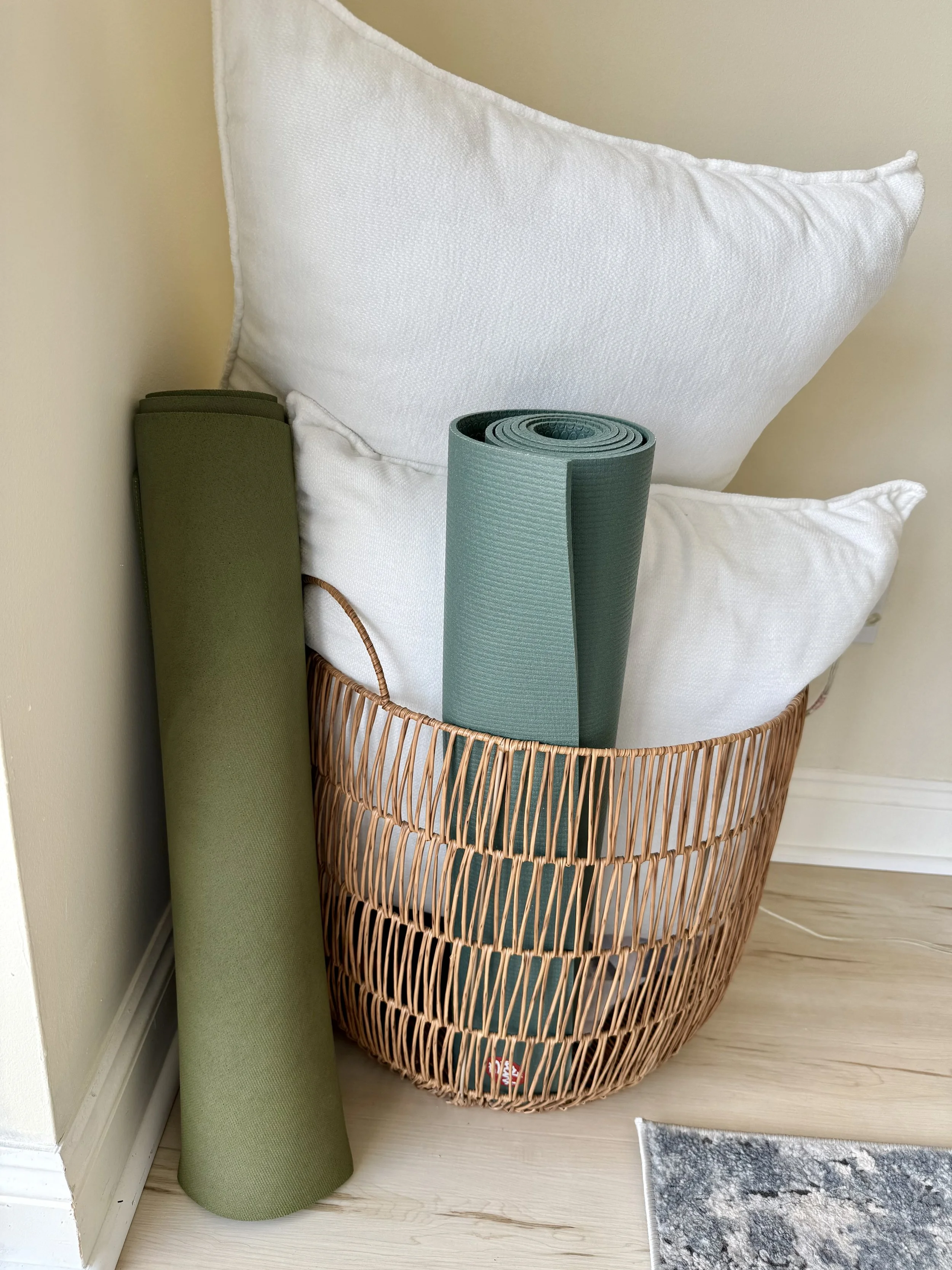 A wicker basket containing two rolled yoga mats, one green and one blue, placed on a light-colored wooden floor next to a white wall, with a small gray and white rug nearby.