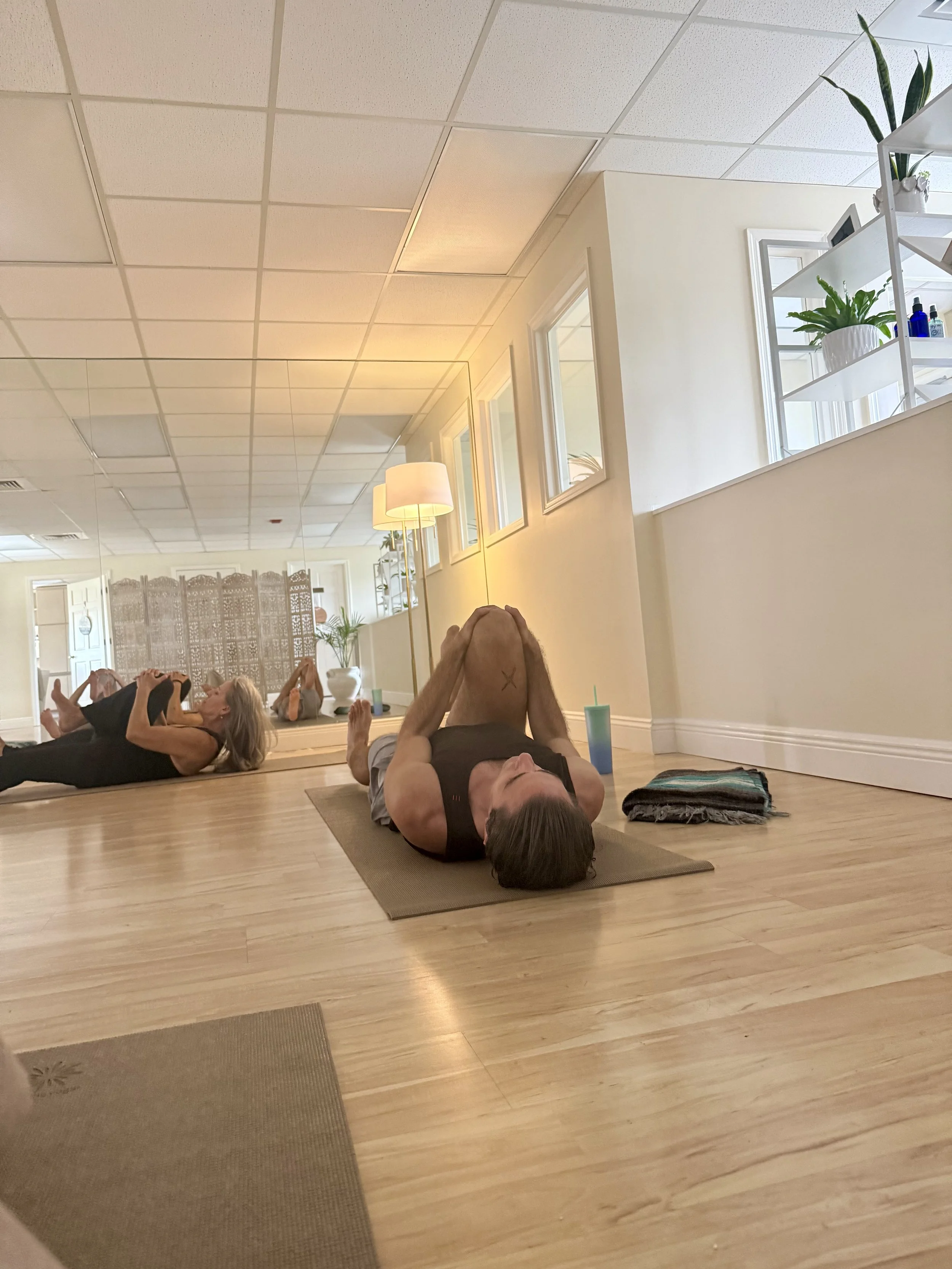 Three elderly women practicing yoga or stretching on mats in a bright, spacious room with wooden floors, mirrors, and plants.