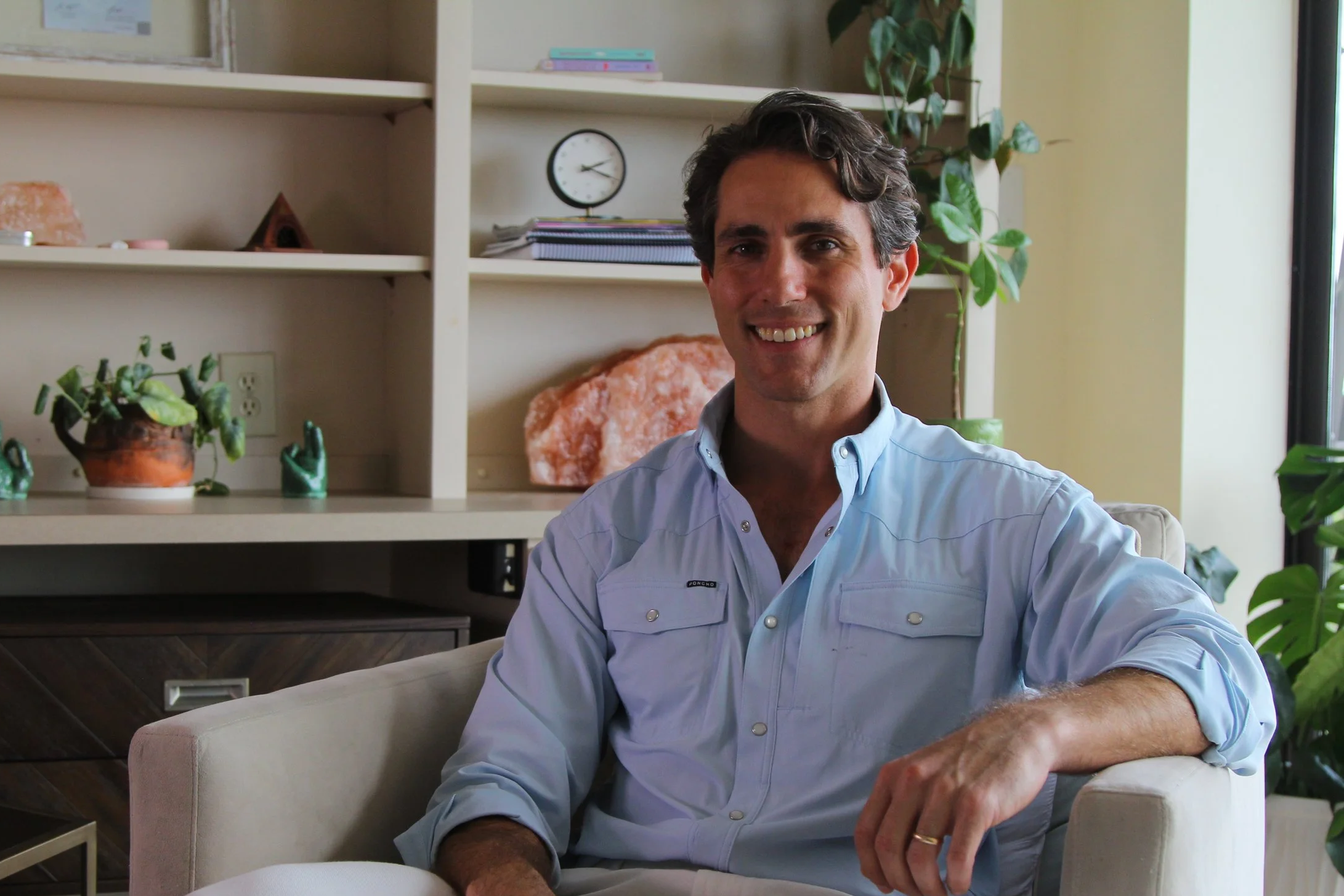 A middle-aged man with dark, curly hair, smiling and sitting in a bright, modern room with shelves, plants, and decorative items.