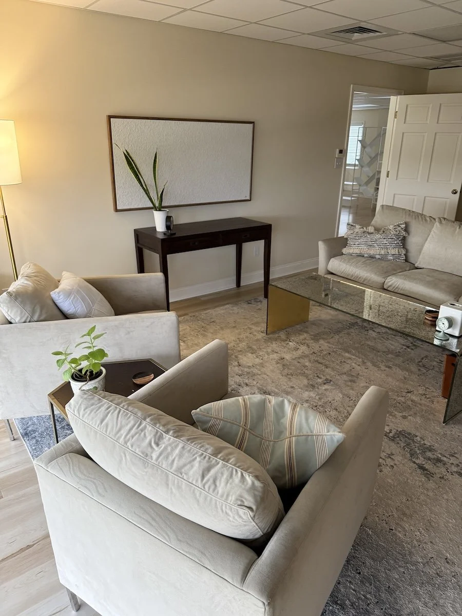 Living room with beige sofas, a glass coffee table, a dark wood console table with a potted plant, a framed empty artwork, and a standing lamp. There is also a doorway leading to another room, and a large area rug.