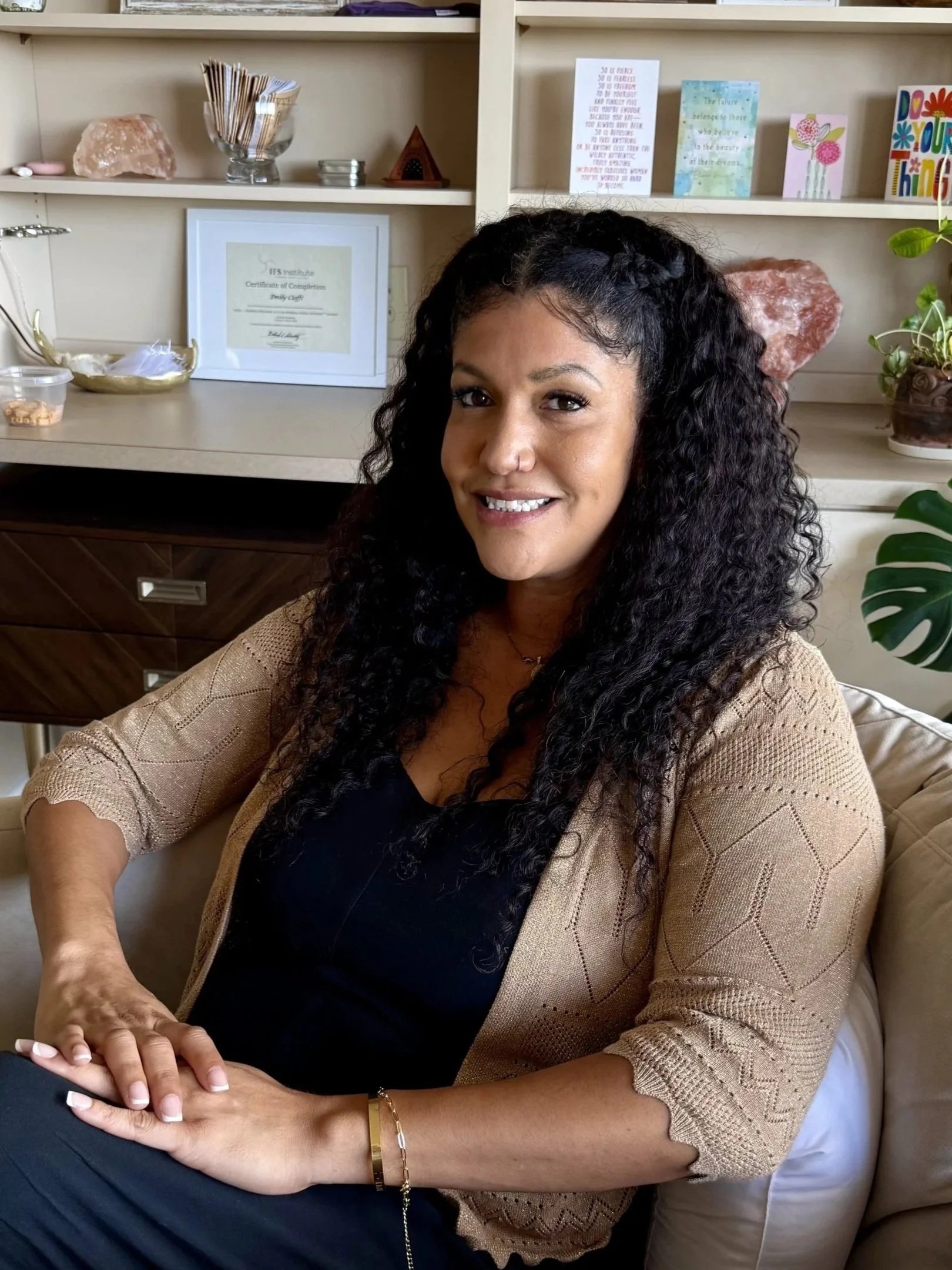 A woman with long, curly black hair sitting on a beige sofa, smiling. She is wearing a black top, a beige knitted cardigan, jewelry, and has manicured nails. In the background, there are shelves with decorative items, a Himalayan salt lamp, a framed certificate, and colorful greeting cards.