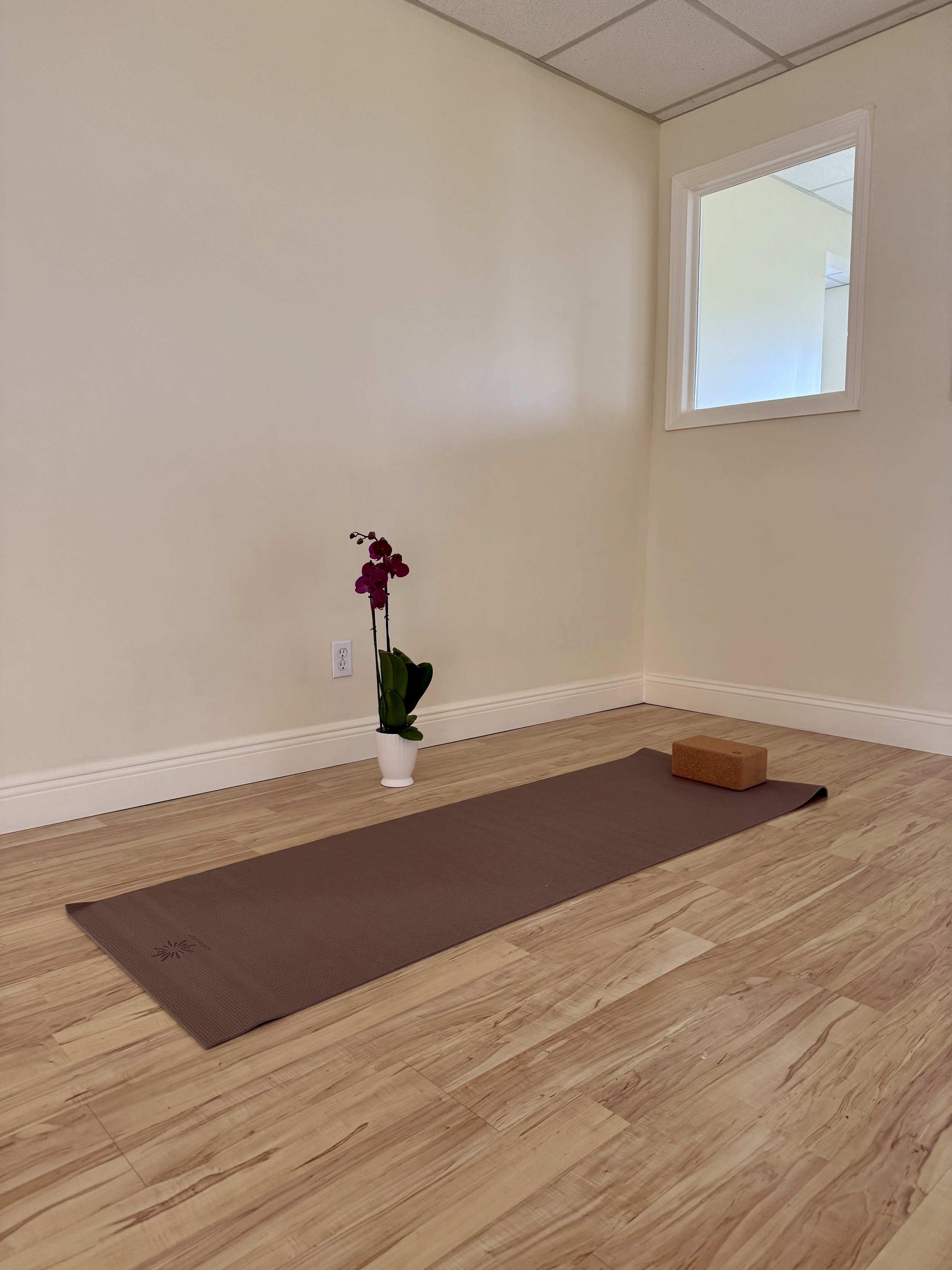 A yoga mat, a brick, and a vase with pink orchids in a room with wooden flooring, cream-colored walls, and a small window.