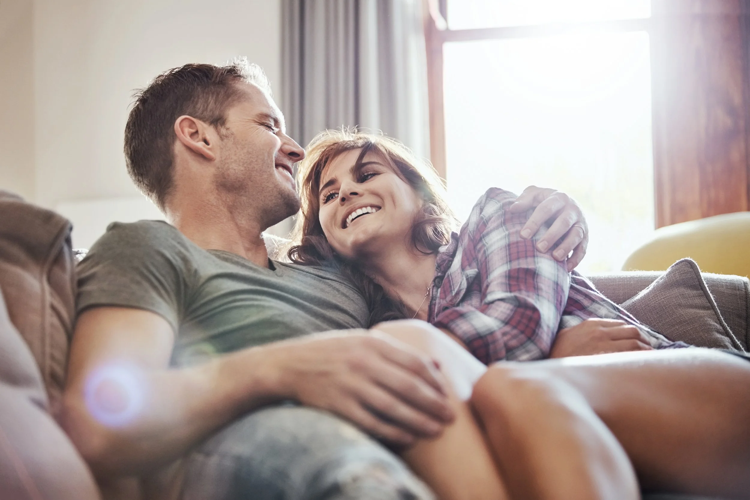 A couple sitting on a couch, smiling, and embracing each other inside a sunlit room with a window.