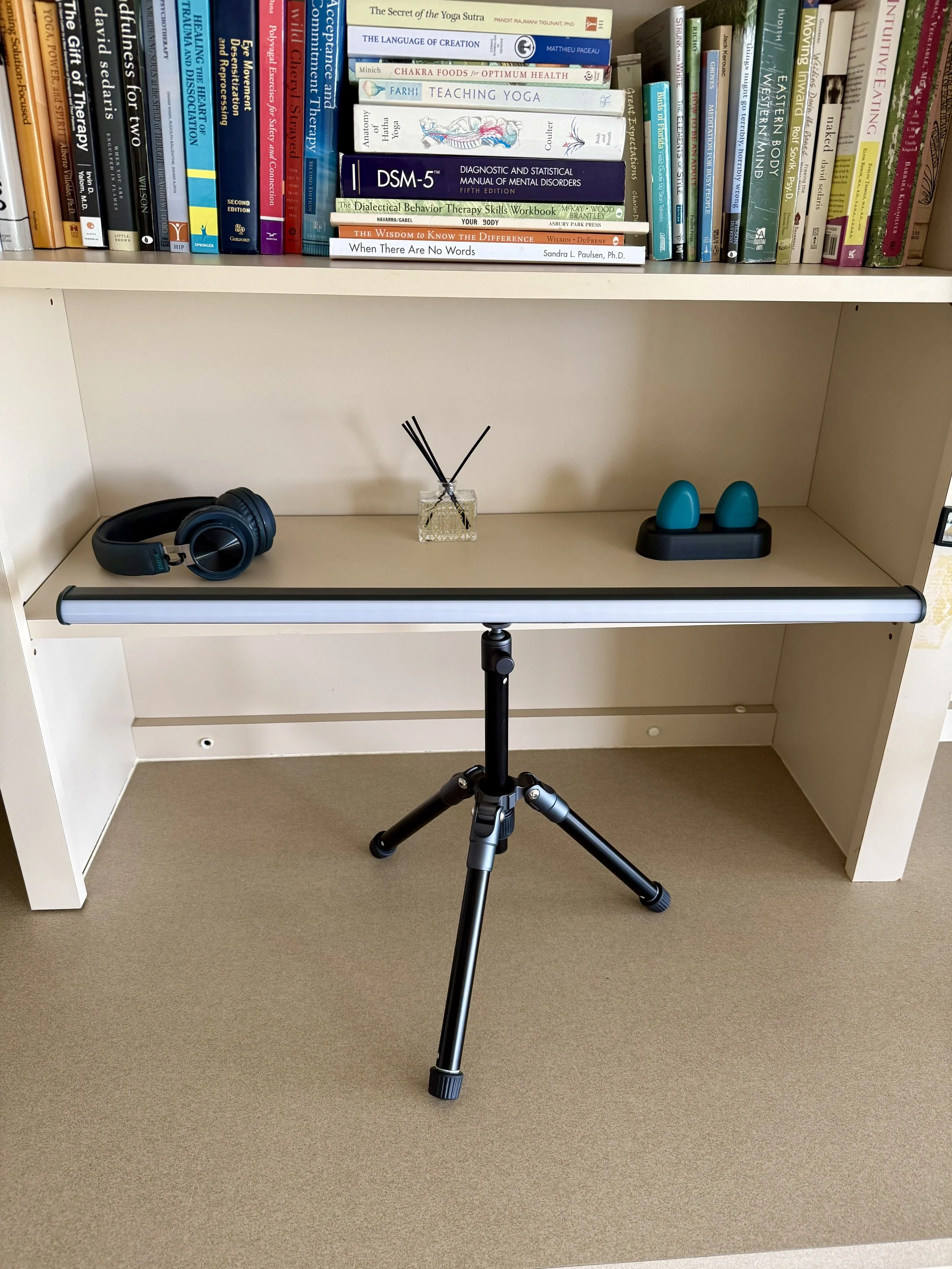 A beige bookshelf with books on mental health, yoga, and therapy, a pair of black headphones, a diffuser with reed sticks, and a black and teal salt lamp, with a black tripod in front.