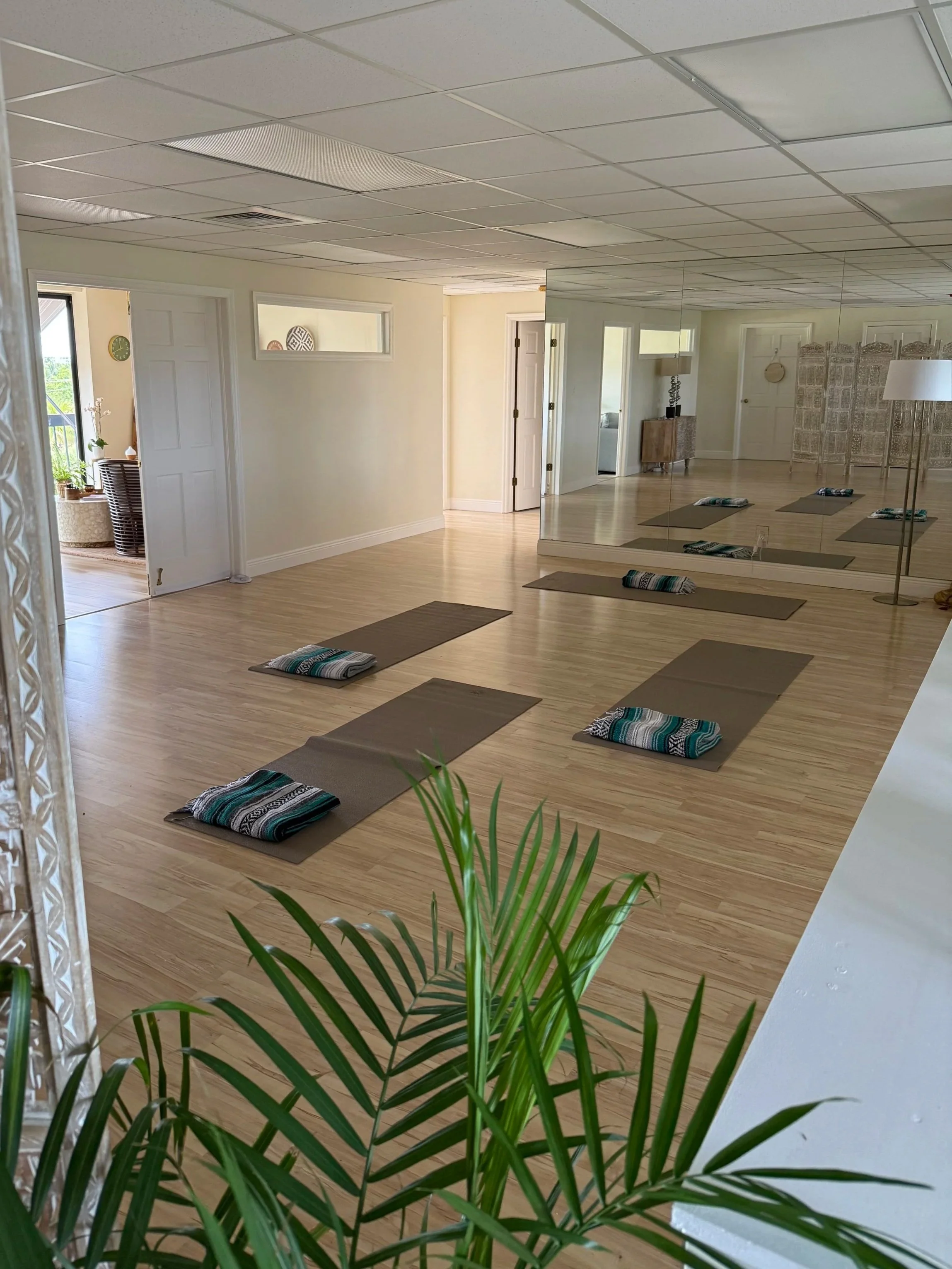 A spacious yoga studio with wooden floors, mirrored wall, and yoga mats with towels arranged on the floor. There is a large mirror on one wall and a lamp, small furniture, and decorative items in the background. A potted plant is in the foreground.