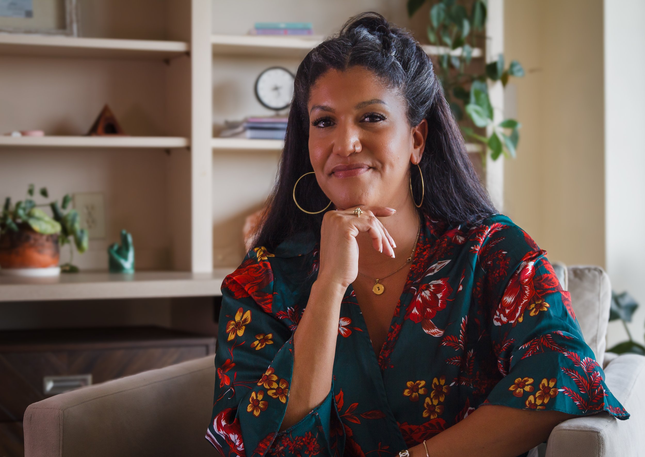 A woman with long dark hair, wearing large gold hoop earrings and a dark floral blouse, sitting indoors with a thoughtful expression, resting her chin on her hand, in front of a bookshelf and plant.