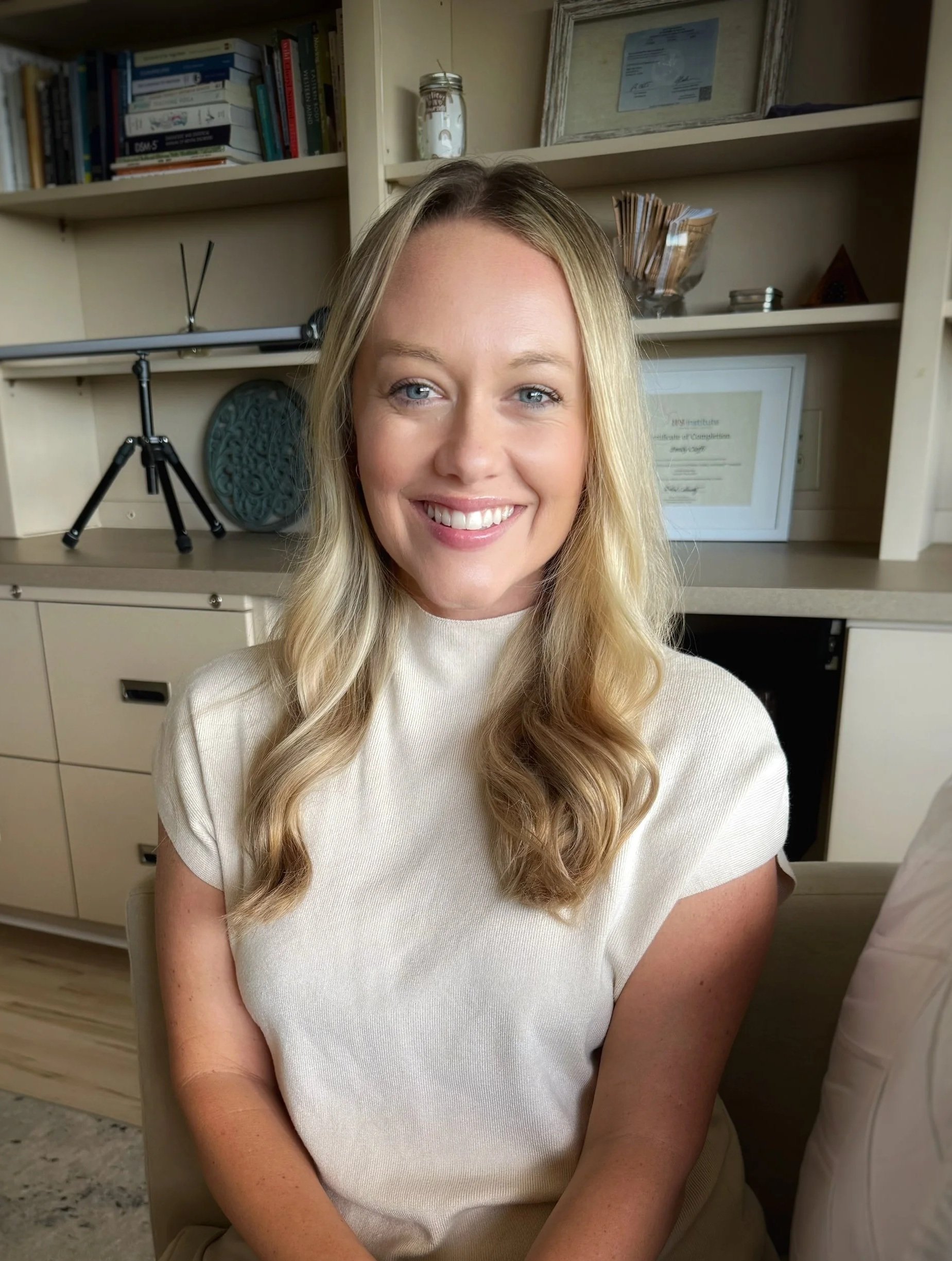 A woman with long blonde hair and blue eyes smiling at the camera, wearing a light-colored top, seated indoors with shelves and framed diplomas or certificates in the background.