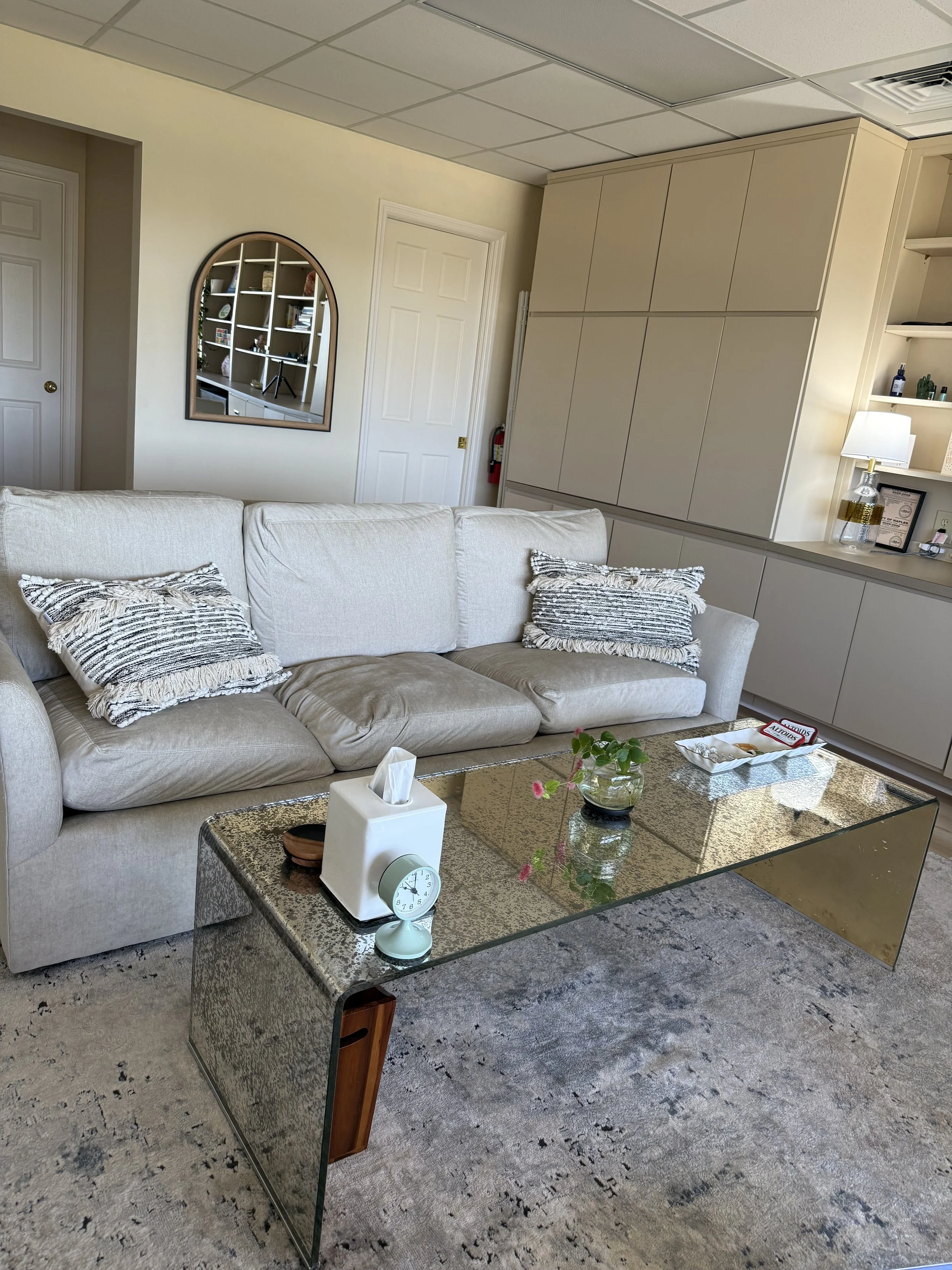 A beige sofa with decorative pillows in a living room, with a glass coffee table, a lamp, and a bookshelf visible in the background.