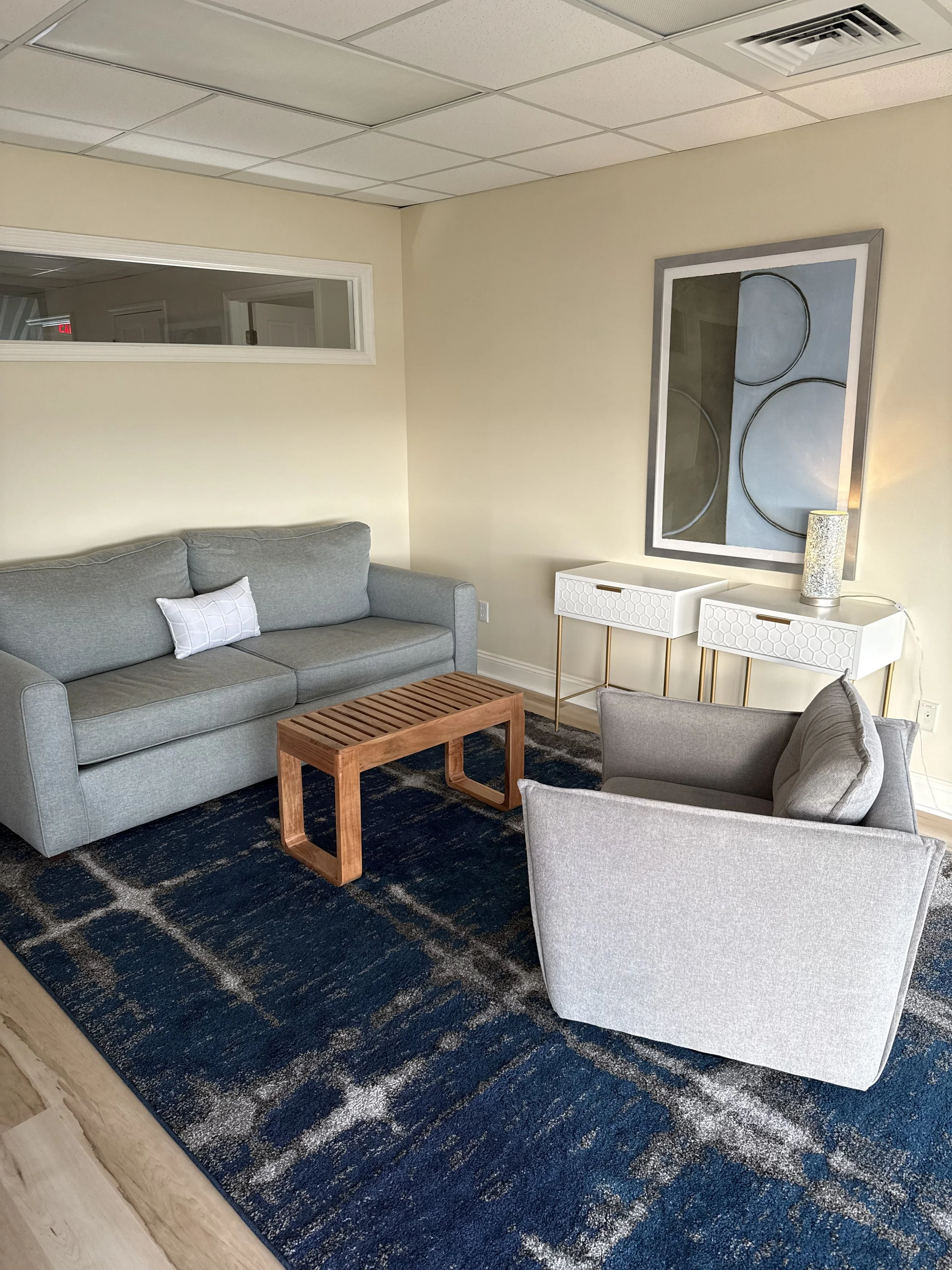 A living room with a gray couch, a wooden coffee table, a gray armchair, a white side table, a framed abstract painting on the wall, and a dark patterned rug.