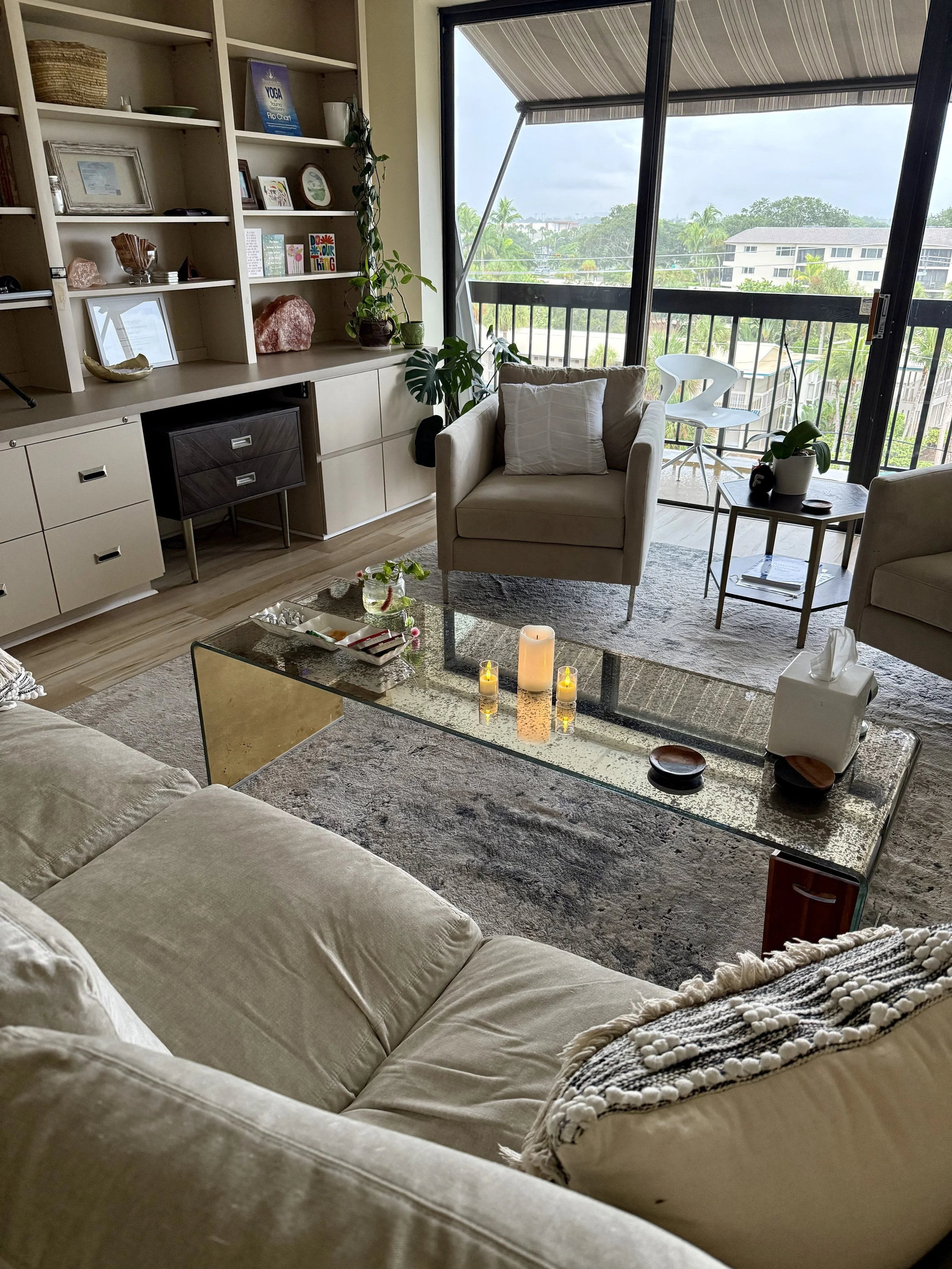 A living room with a beige couch, a glass coffee table with candles, and a fireplace. A bookshelf with decorative items and books is against the wall. A sliding glass door opens to a balcony with outdoor chairs and a view of trees and apartment buildings.