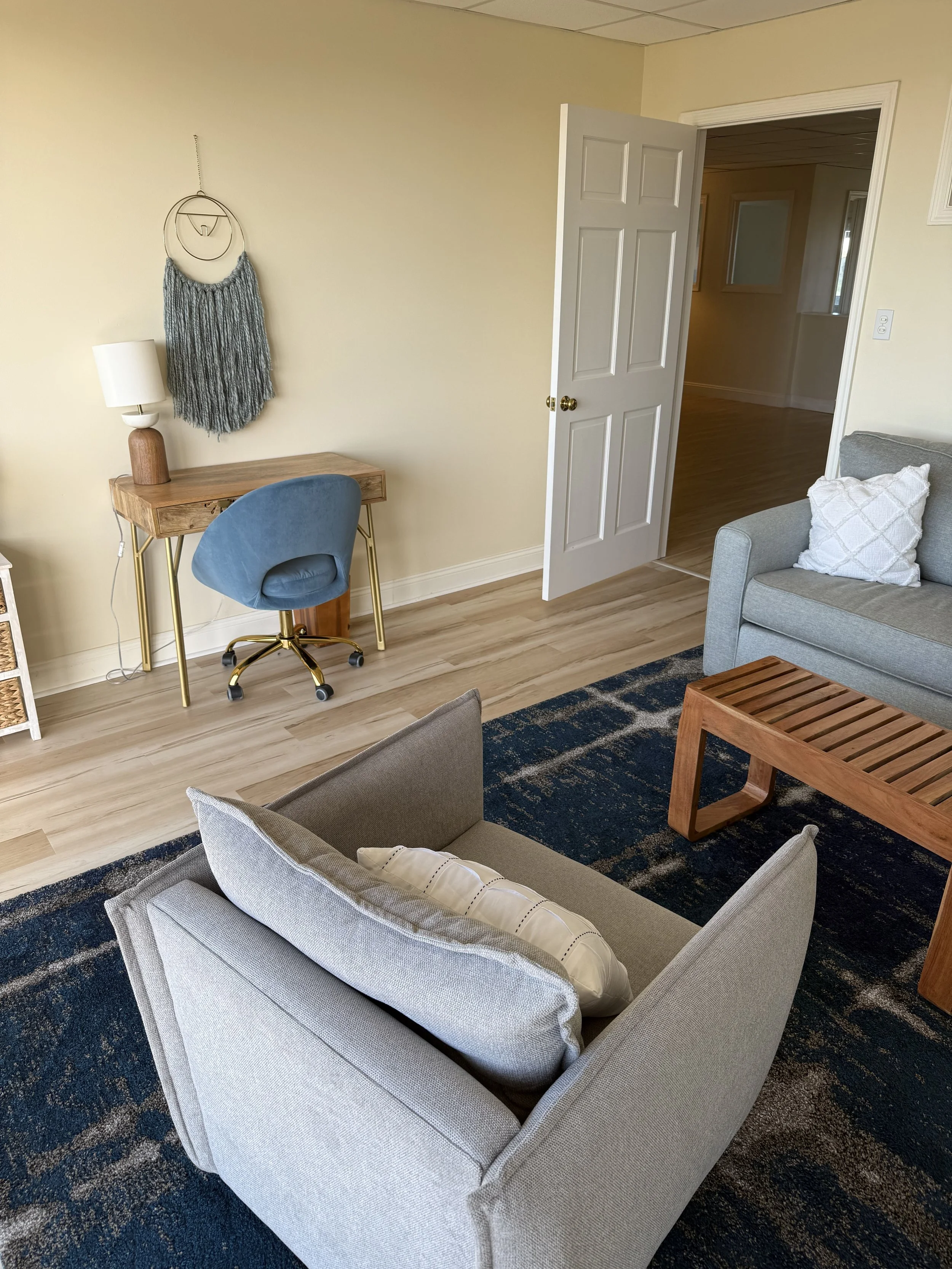 A cozy living room with a beige sofa, decorative pillows, a wooden coffee table, a blue accent chair at a small desk with a modern lamp, a textured wall hanging, and a dark patterned rug, with an open door leading to a hallway.