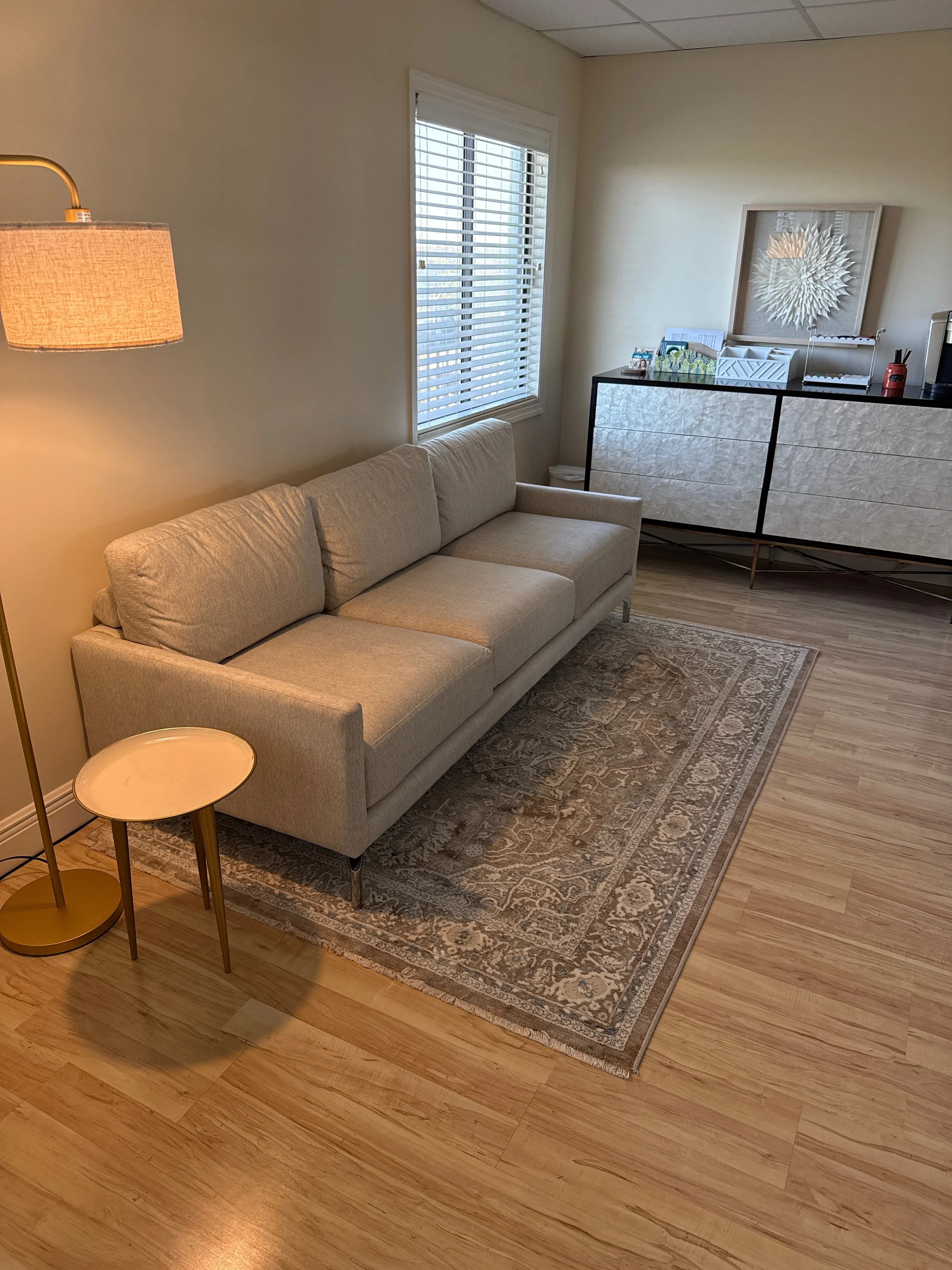 A beige sofa with metal legs situated on a patterned rug in a room with hardwood floors, a window with blinds, a chest of drawers with decorative items on top, and a floor lamp beside the sofa.