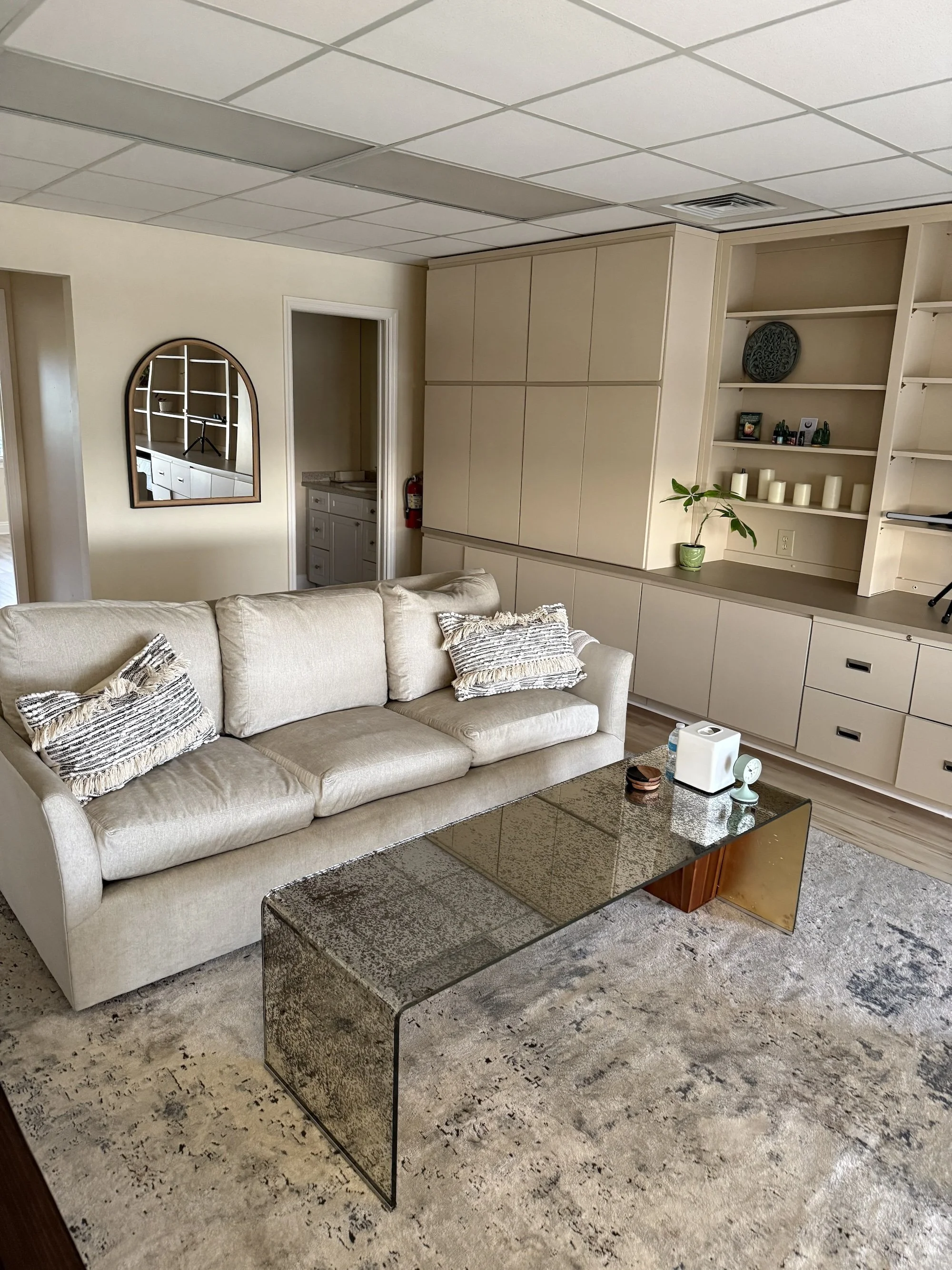 Living room with beige sofa, black granite coffee table, built-in shelves with candles and decorations, potted plant, mirror, and doorway, in an office or home interior.