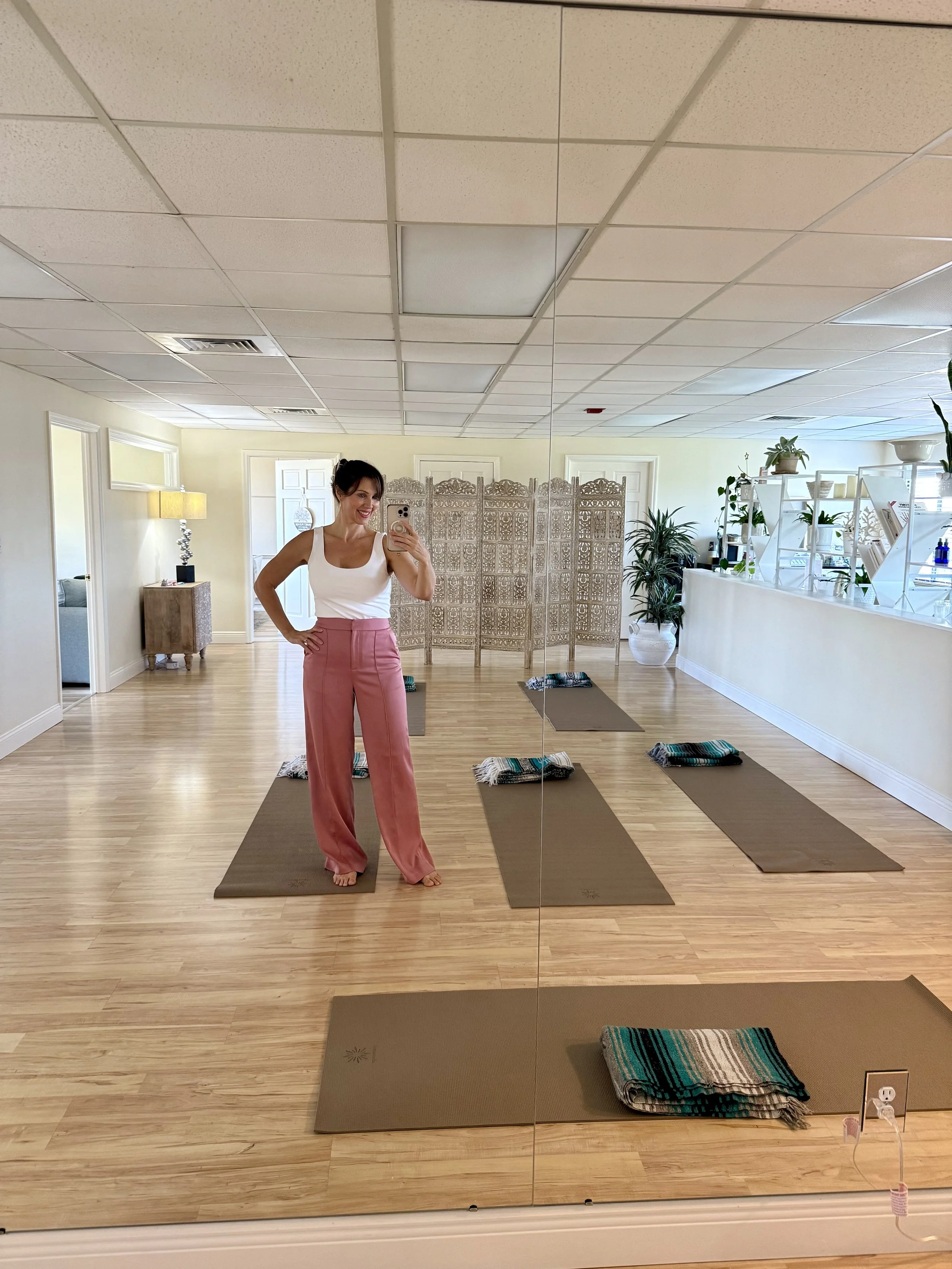 a woman standing in a yoga studio in front of a large mirror, taking a selfie, wearing a white tank top and pink wide-leg pants, with yoga mats and a folded blanket on the floor, and a folding screen and plants in the background.