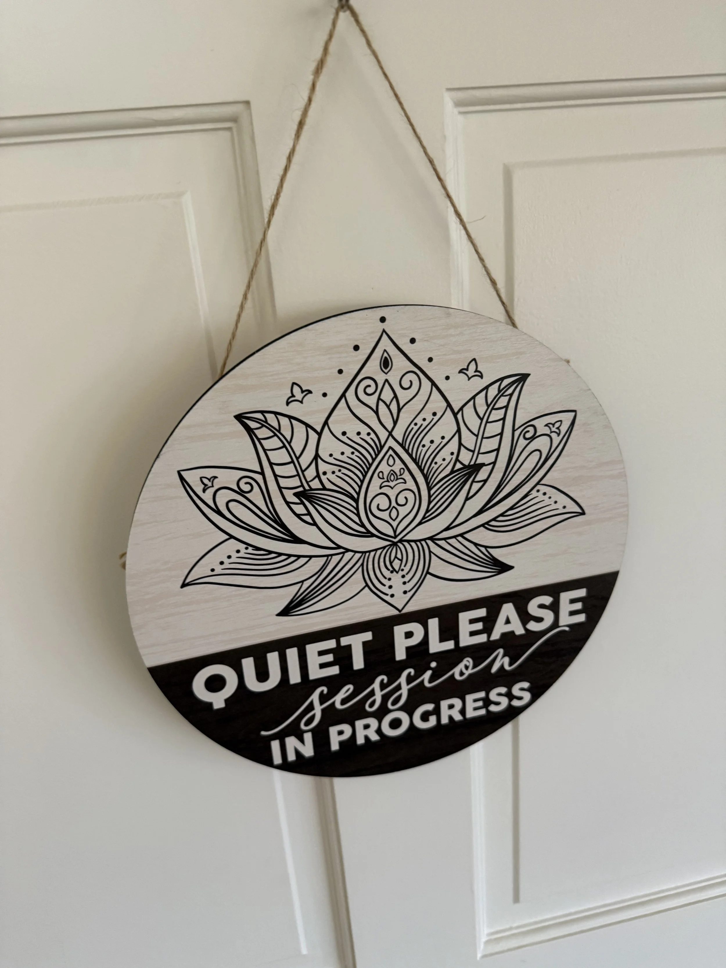 A circular sign hanging from a wall with a rope, featuring an intricate mandala design in black and white, with the text 'Quiet Please' and additional words partially visible.