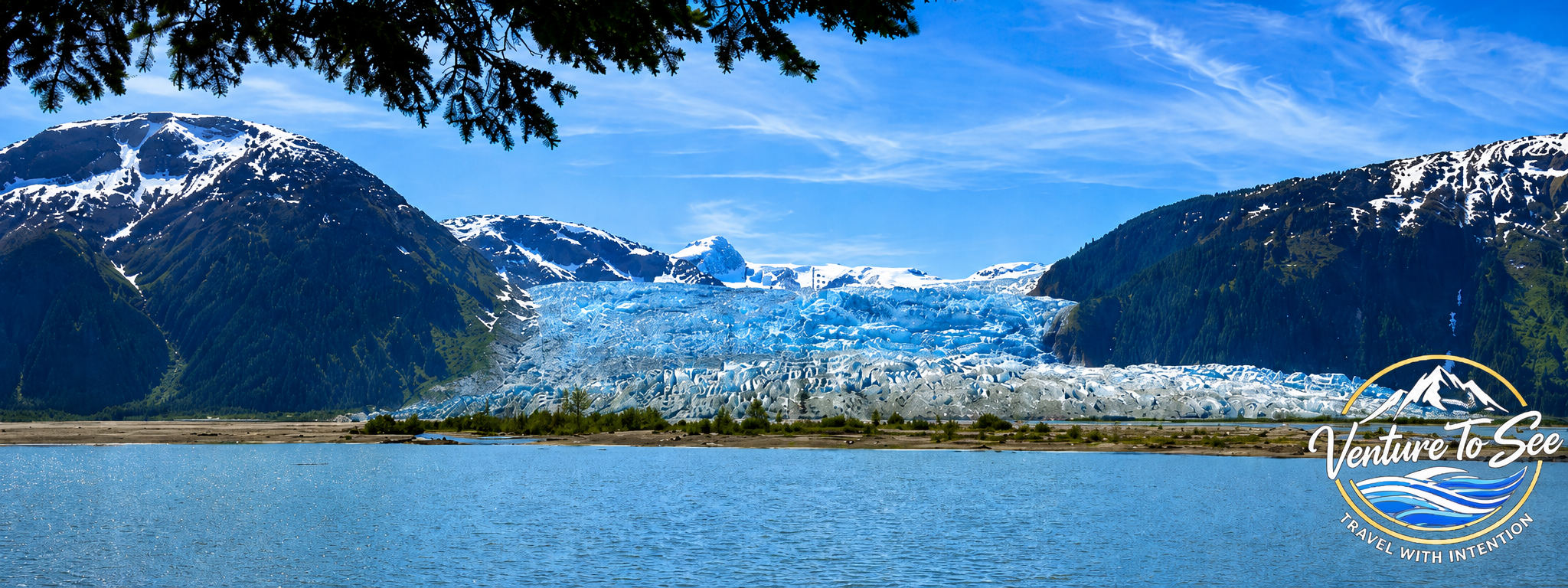 A glacier nestled between mountain ranges with snow and ice, located near a body of water under a clear blue sky, with some trees in the foreground.