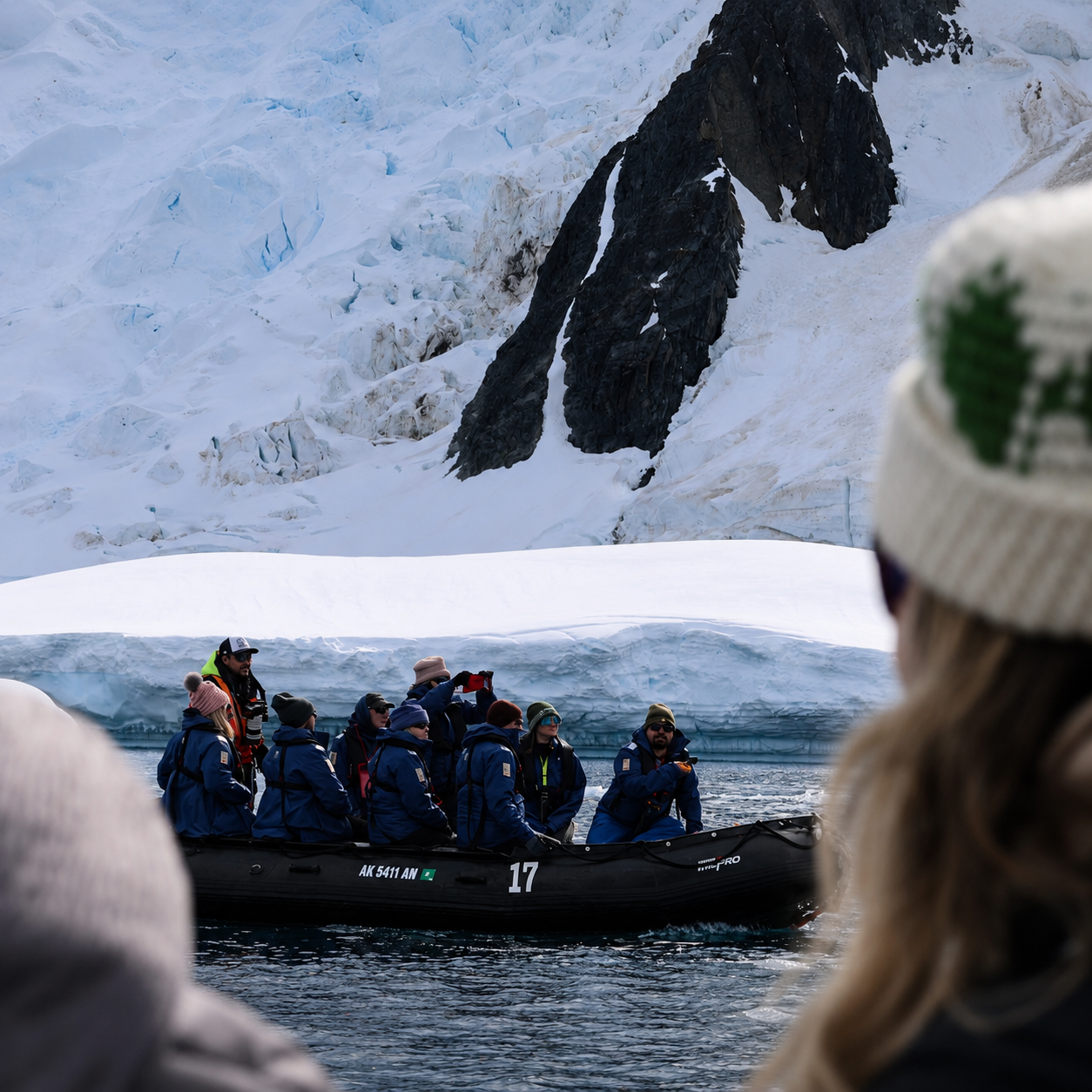 A group of people in blue jackets riding in a black inflatable boat near an icy shoreline with large icebergs and snow-covered mountains in the background.