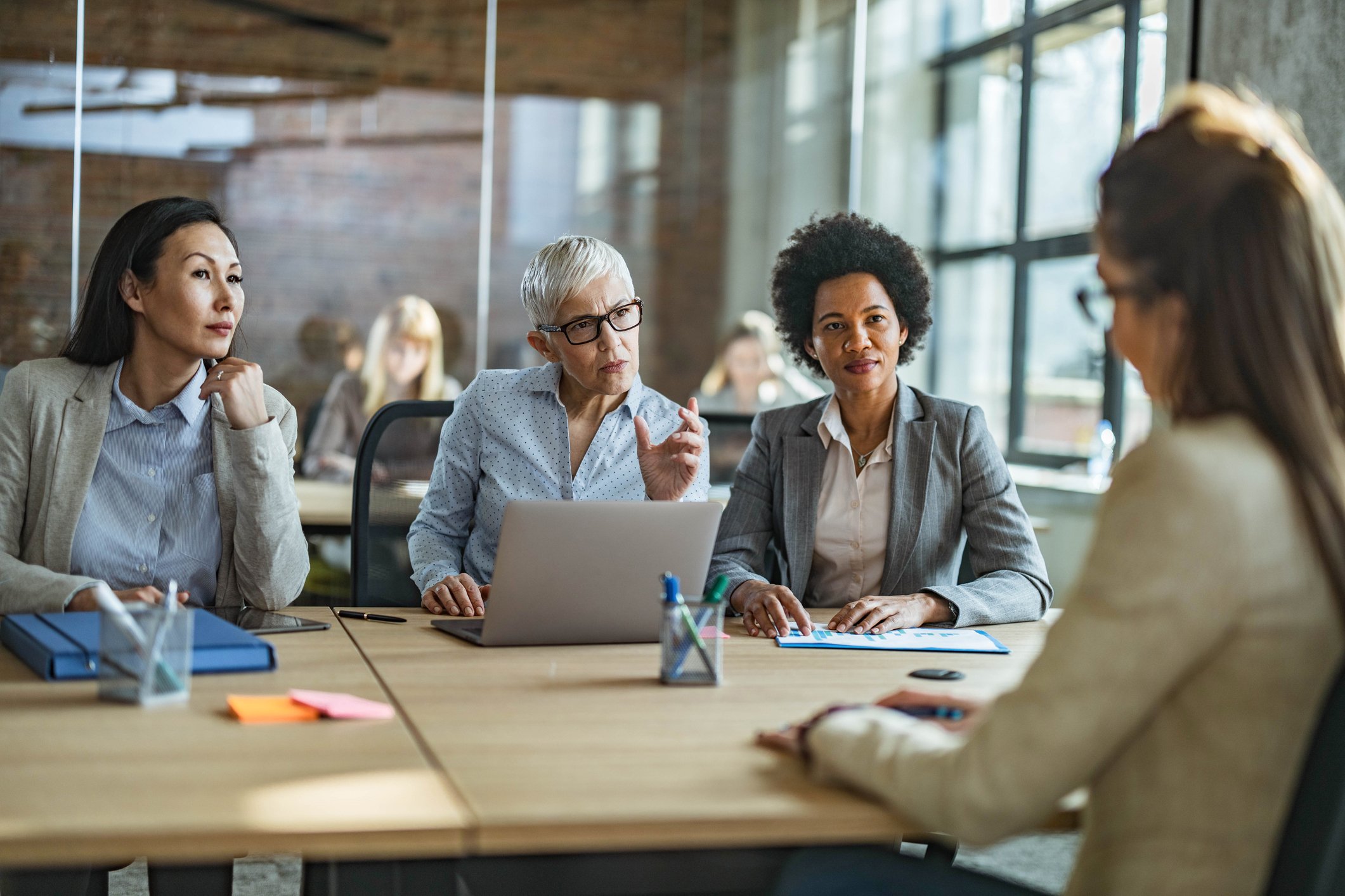 A diverse group of women engaged in a serious discussion at a business meeting in a modern office conference room.