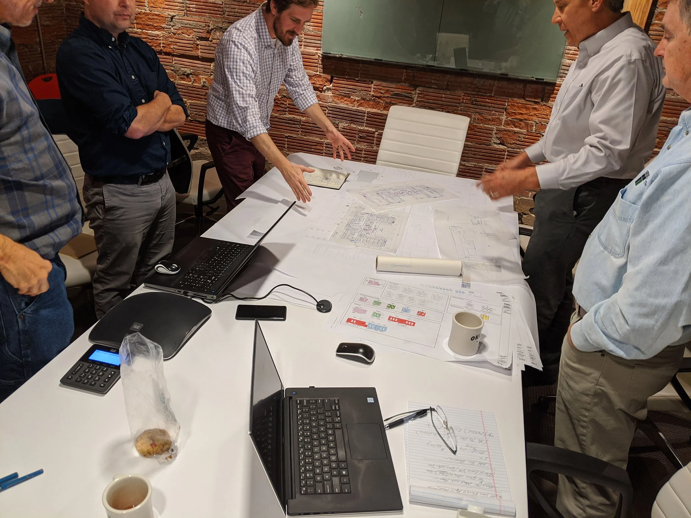 A group of five people is gathered around a table with architectural blueprints, documents, laptops, and office supplies, engaged in discussion in a room.