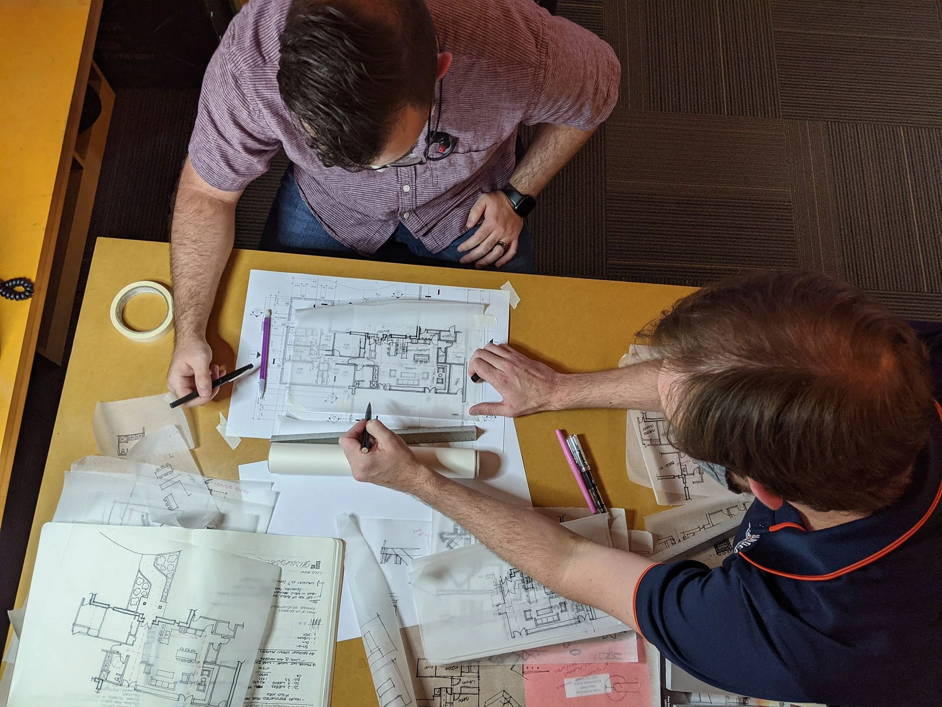 Two individuals are working on architectural blueprints and floor plans at a yellow table, surrounded by various papers and drafting tools.
