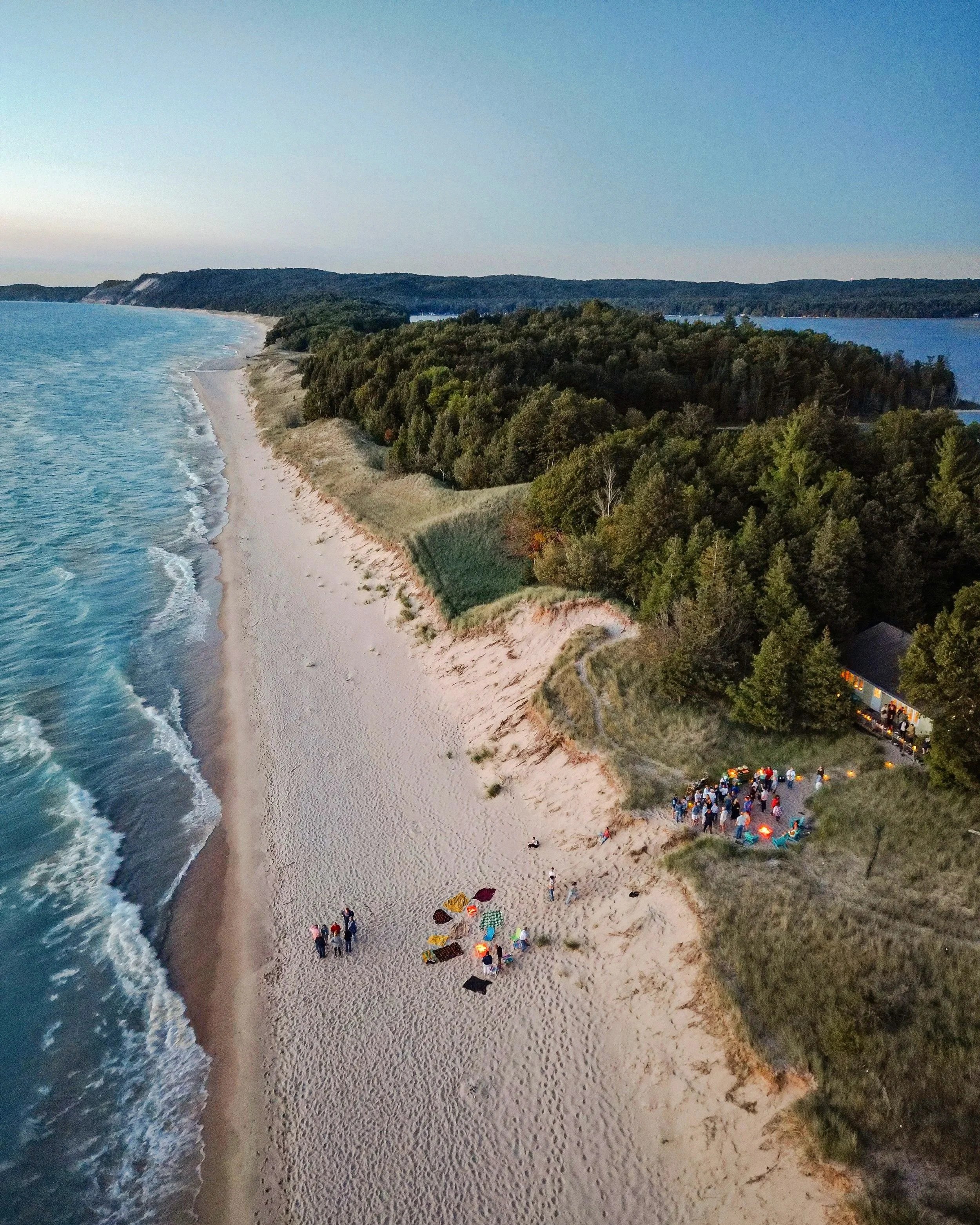 A beach at sunset with people gathered around a bonfire near the water, surrounded by trees and dunes.