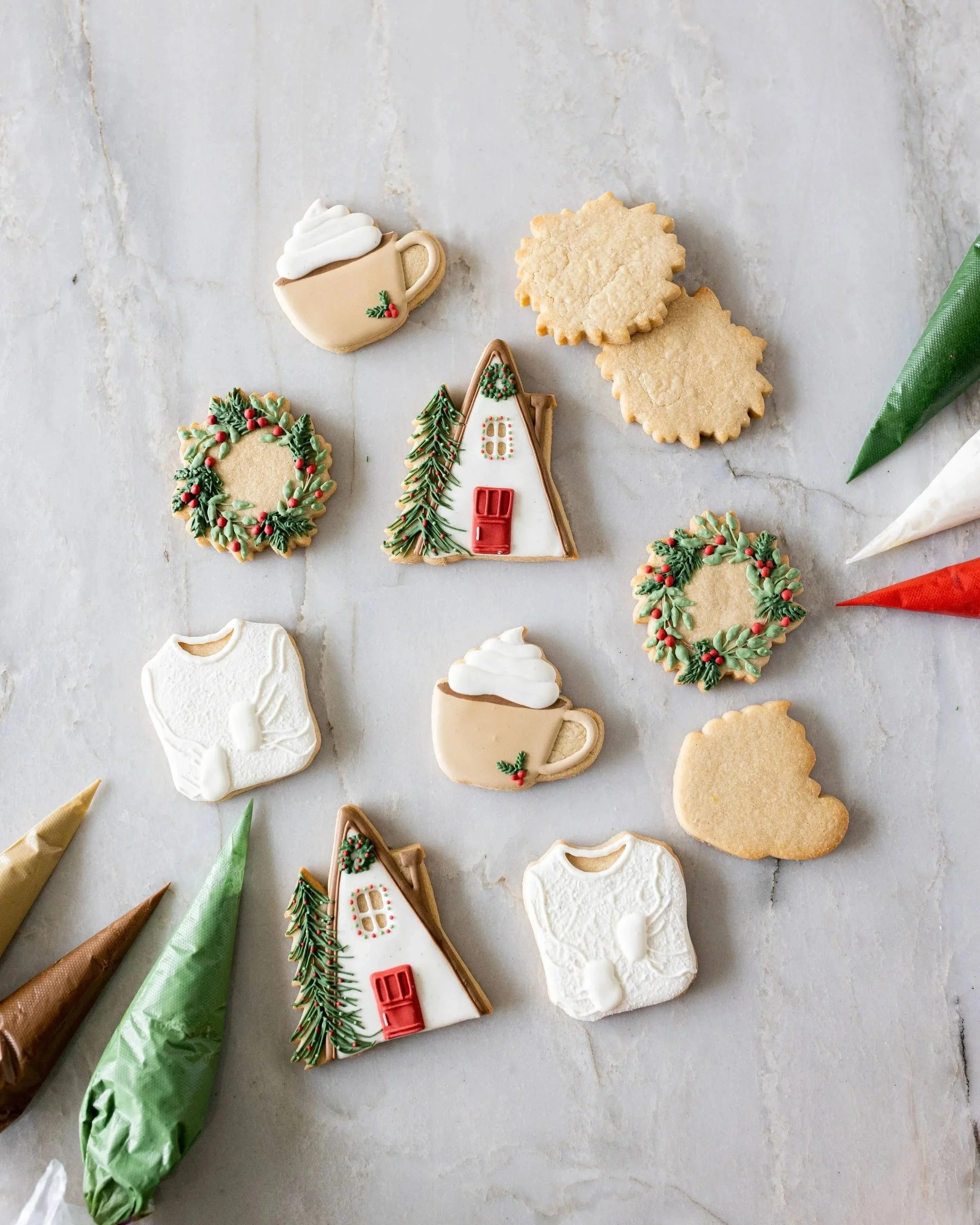 Decorated Christmas cookies on a white surface, including a house with a Christmas tree, two round cookies with a scalloped edge, and a wreath. There are also frosting piping bags in green, gold, and brown on the side.